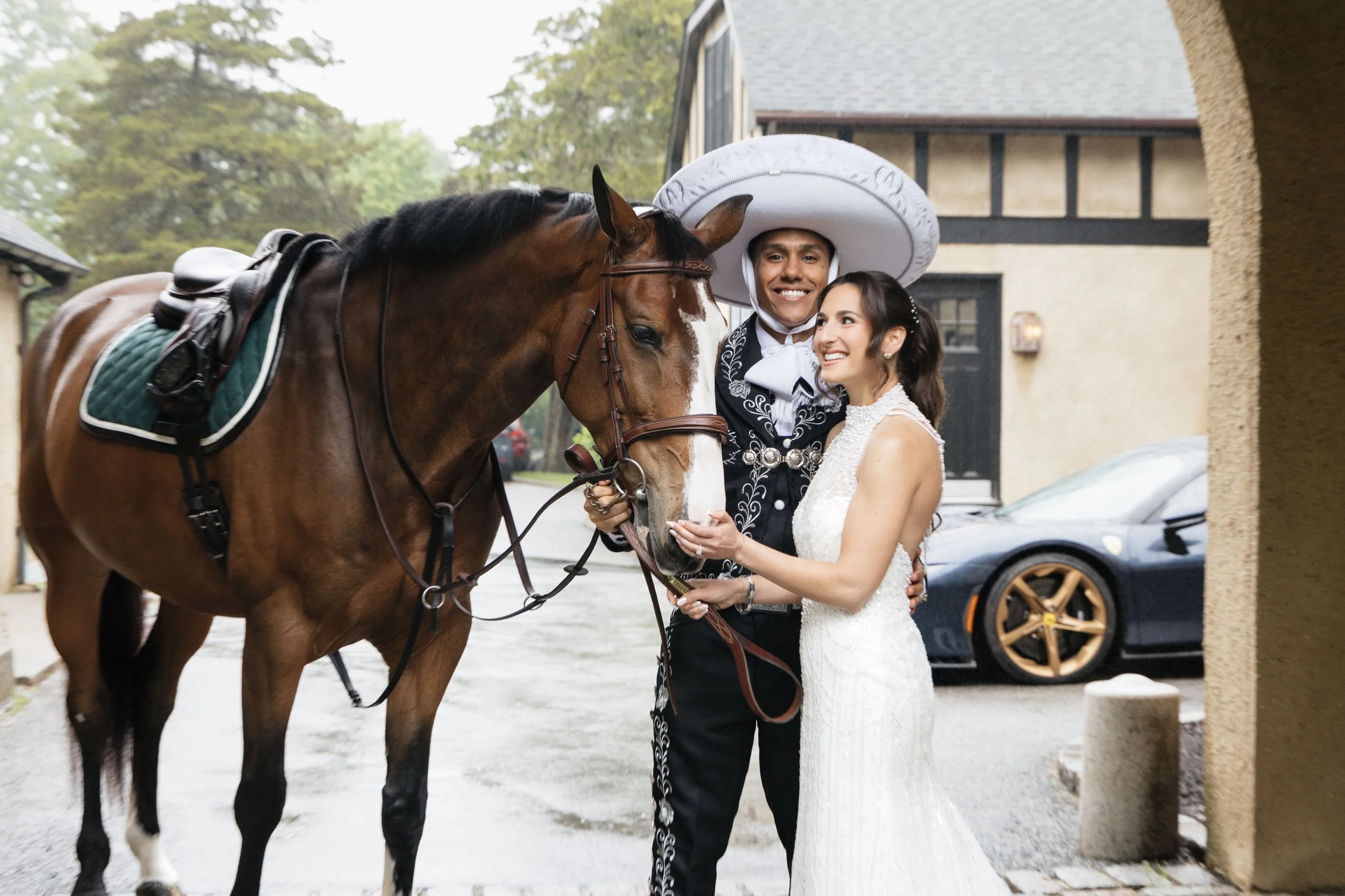 A bride and groom smiling and holding a horse's reins outside a building, with trees and a car in the background.