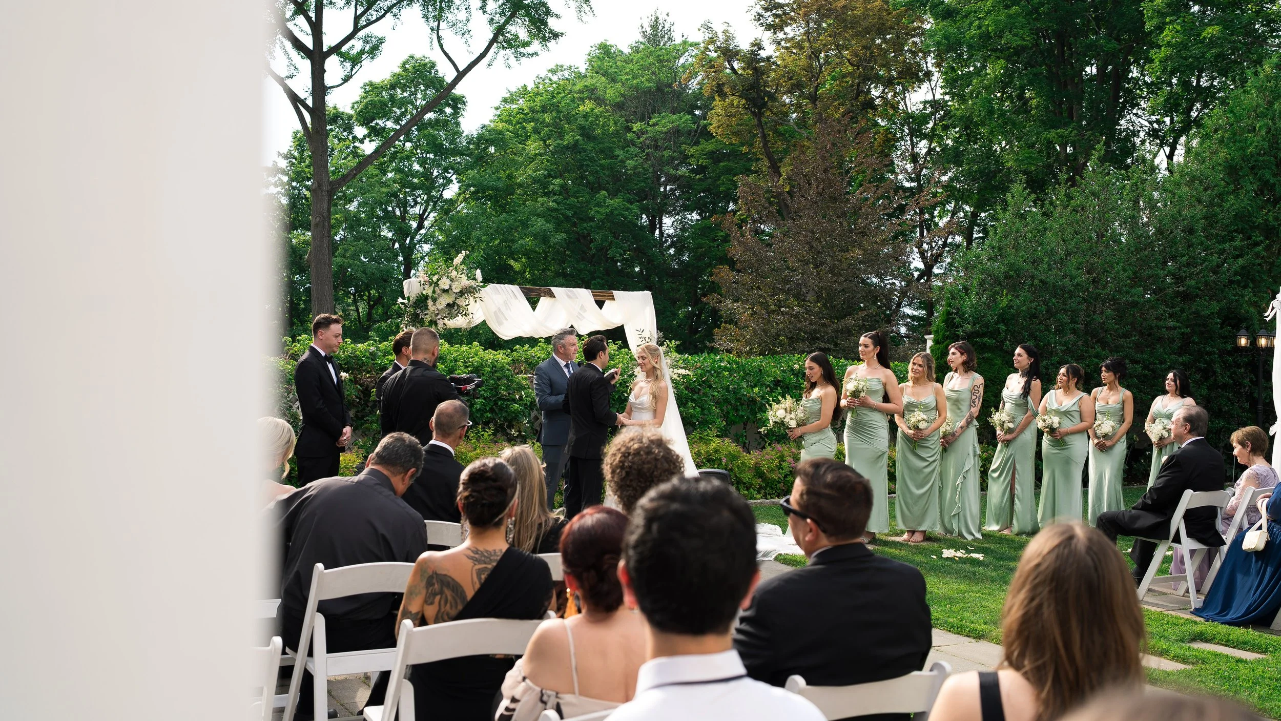 A wedding ceremony taking place outdoors with the bride and groom exchanging vows, surrounded by bridesmaids in light green dresses holding bouquets, and guests seated on white chairs, set against lush green trees and a decorated arch.