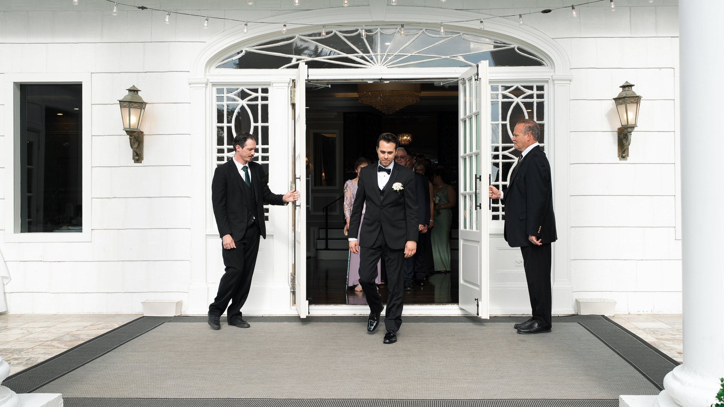 A groom in a black tuxedo walking out of a building with two men holding the door open on either side. People are inside the building, and there are decorative lights and wall sconces on the exterior.