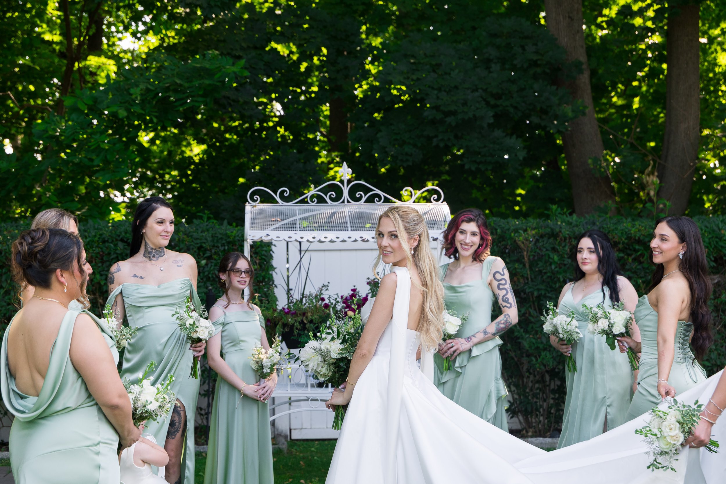 A group of women in sage green bridesmaid dresses and a bride in a white wedding gown gathered outdoors in a garden with lush green trees, holding bouquets of white flowers during a wedding celebration.