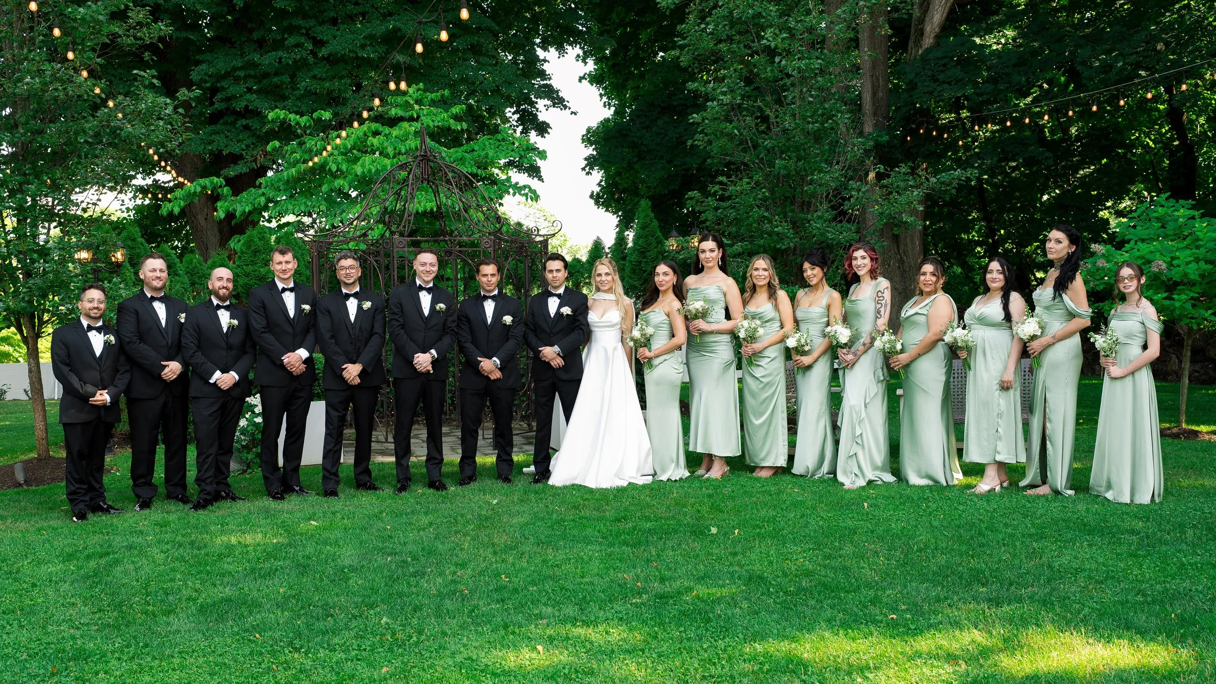 A wedding party of fifteen people standing outdoors on a grassy field with green trees and string lights overhead. The bride and groom are in the center, with the bride wearing a white gown and the groom in a black tuxedo. Bridesmaids in matching lig