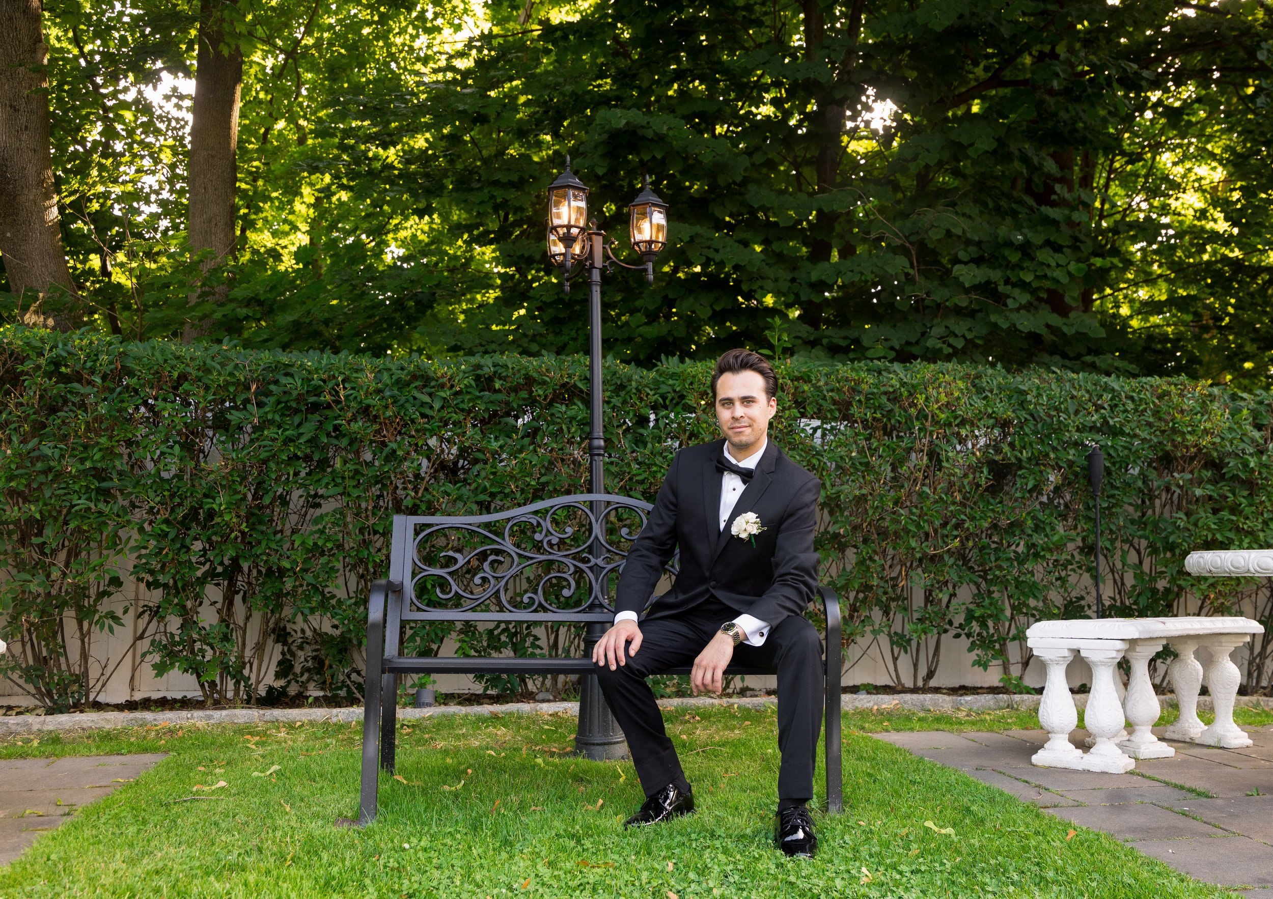 Man in a black tuxedo sitting on a decorative metal bench outdoors, surrounded by lush green foliage and trees, with a vintage-style street lamp behind him.
