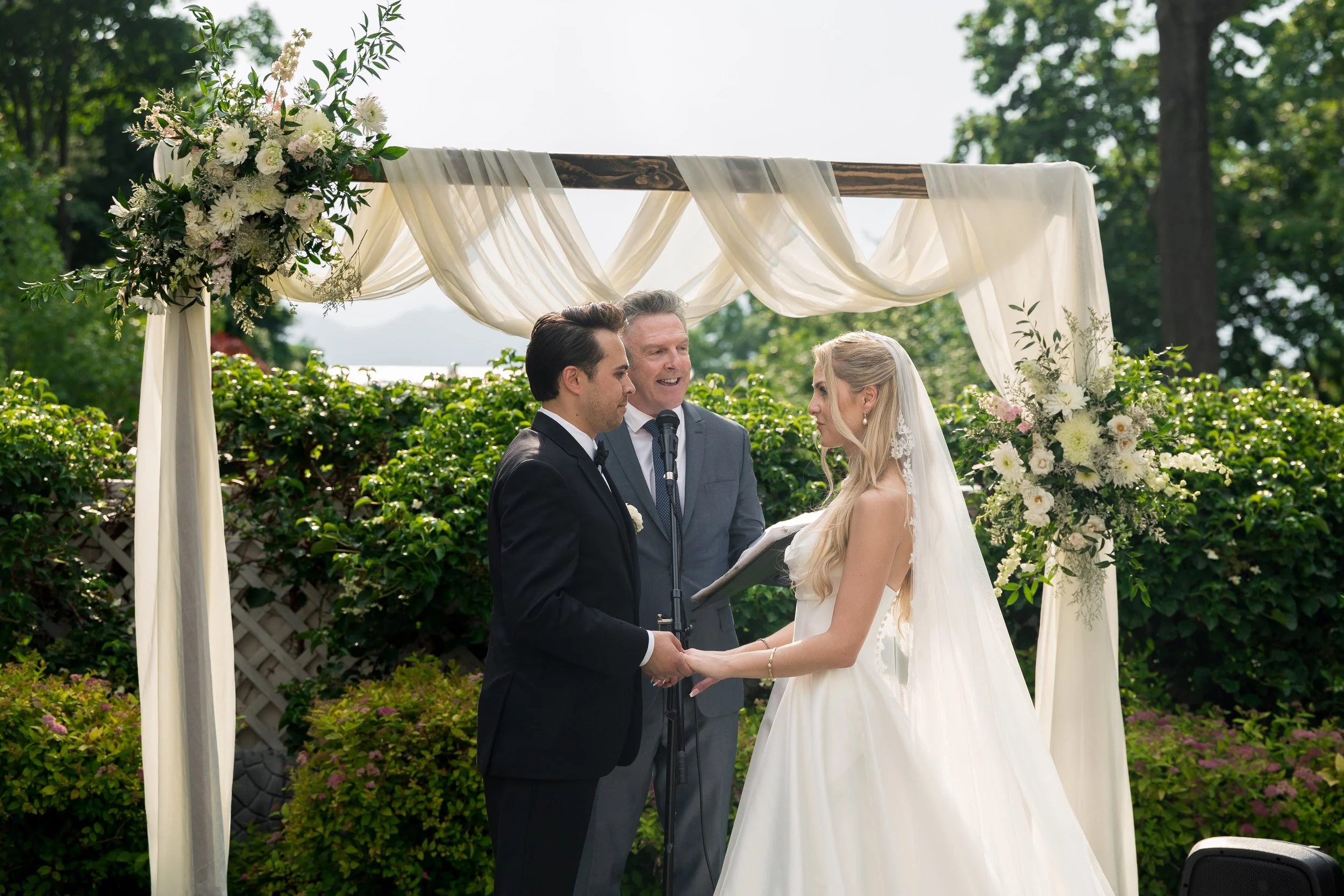 A couple getting married outdoors under a wooden arch decorated with white flowers and sheer fabric, with an officiant standing between them and holding a microphone.