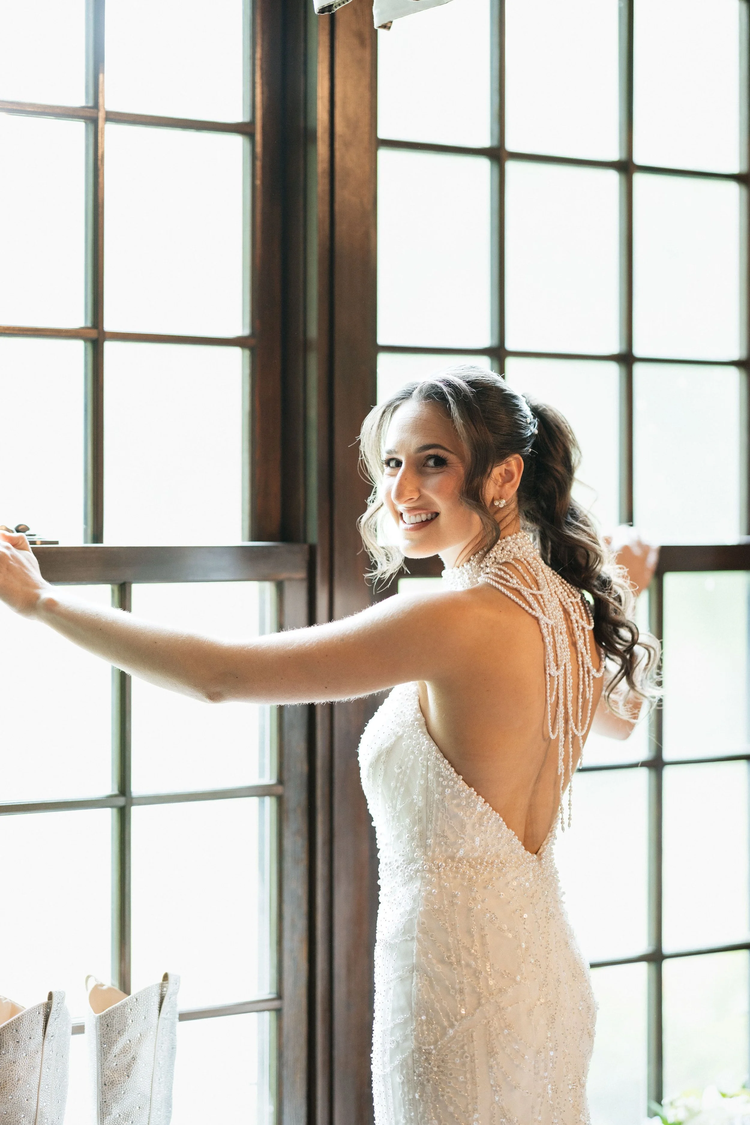 A bride with a big smile, wearing a pearl-adorned wedding dress, standing near a large window with wooden framing, with her right hand extended outward.