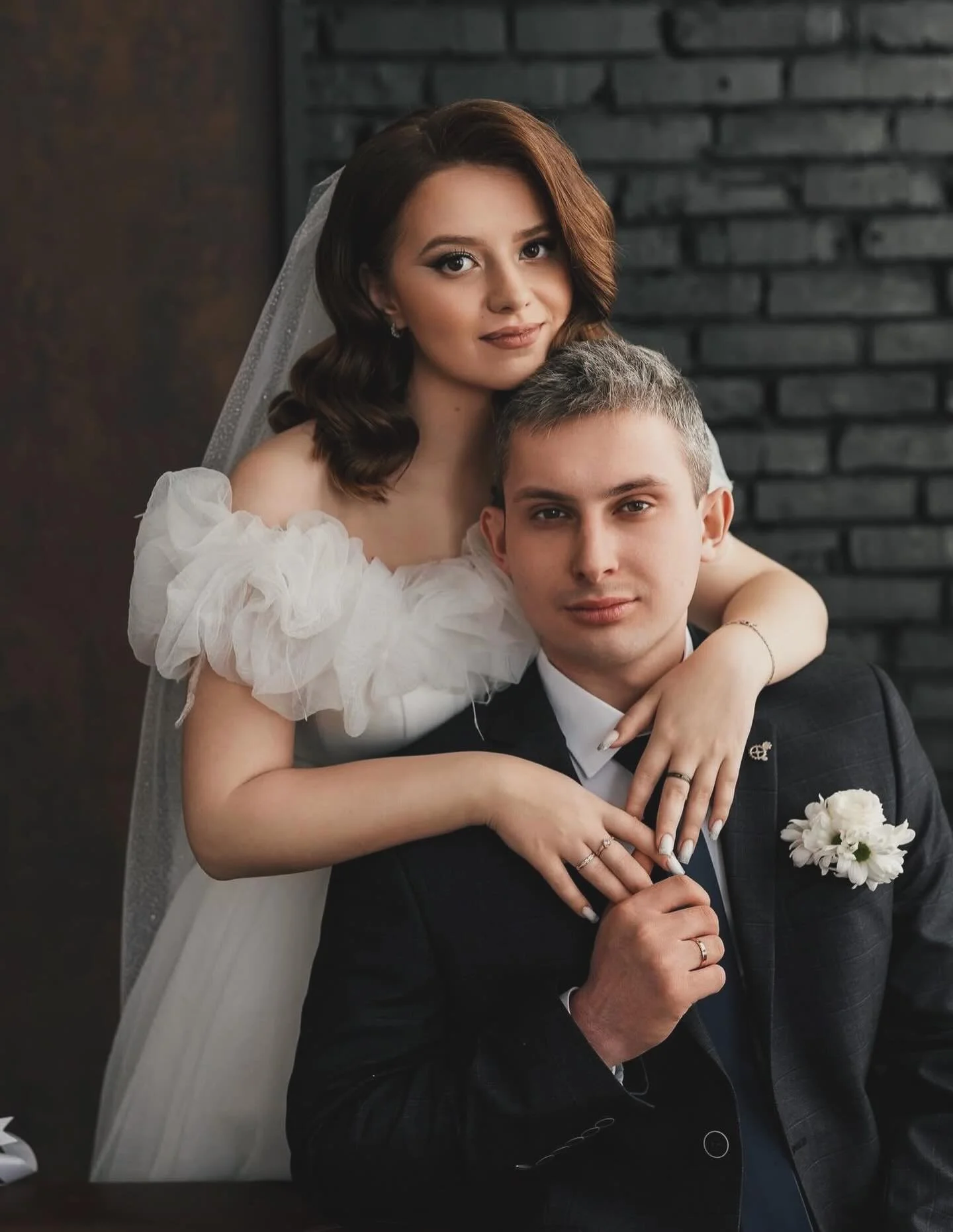A bride with brown hair, wearing a veil and white dress, and a groom with short gray hair, wearing a black suit and white shirt, are posing together indoors in front of a dark brick wall. The bride is resting her arm on the groom's shoulder and both are looking at the camera, with the bride smiling softly and the groom having a neutral expression.