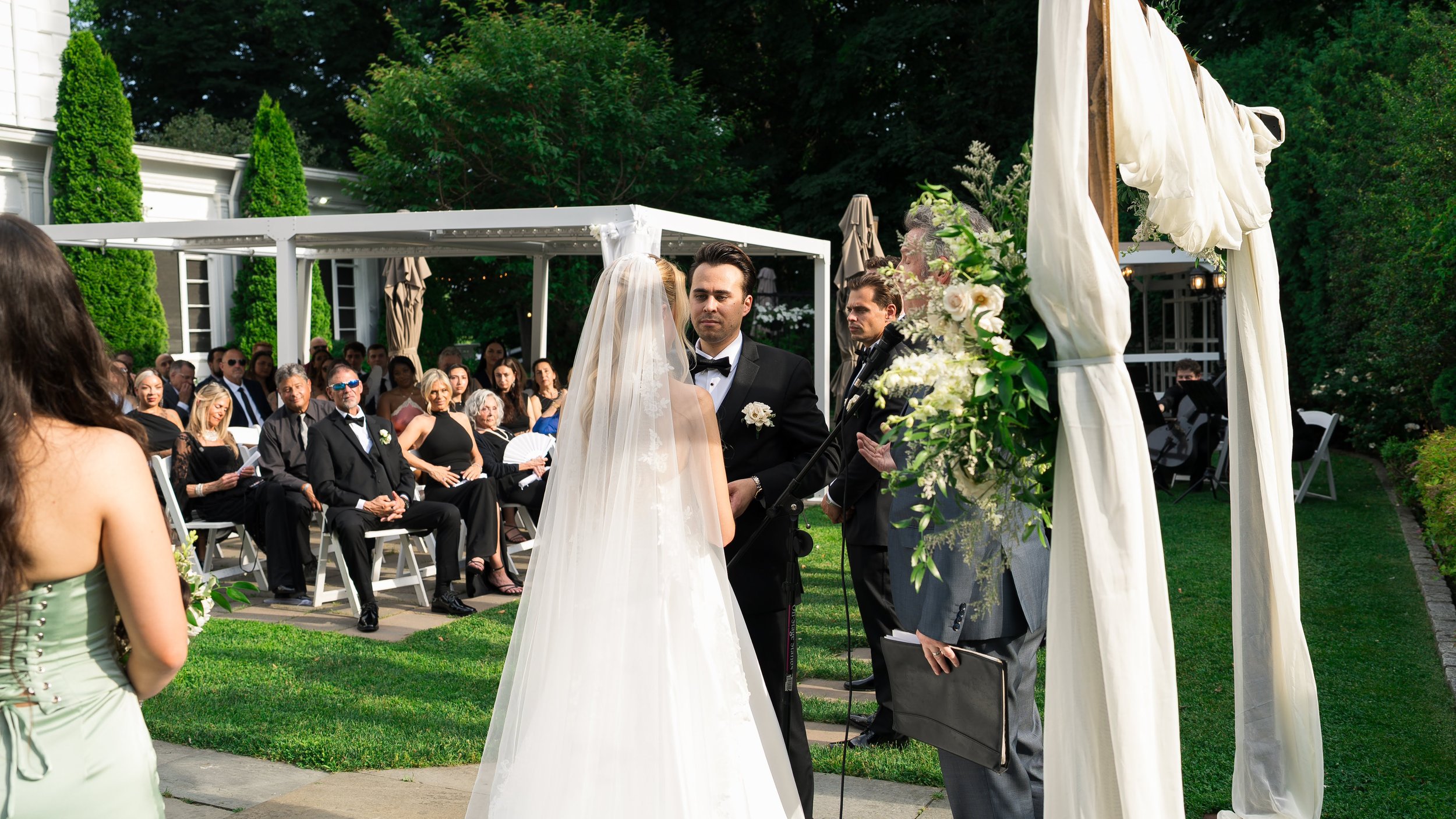 A couple standing at a wedding altar outdoors with guests seated in the background. The scene features lush greenery and a white wedding canopy decorated with flowers.