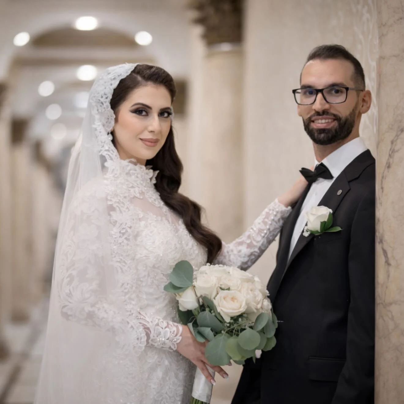 Bride and groom in wedding attire posing in an elegant hallway. The bride wears a lace wedding dress, a lace veil, and holds a bouquet of white roses and greenery. The groom wears a black tuxedo with a white shirt, black bow tie, and a white boutonniere. They are smiling and looking at the camera.