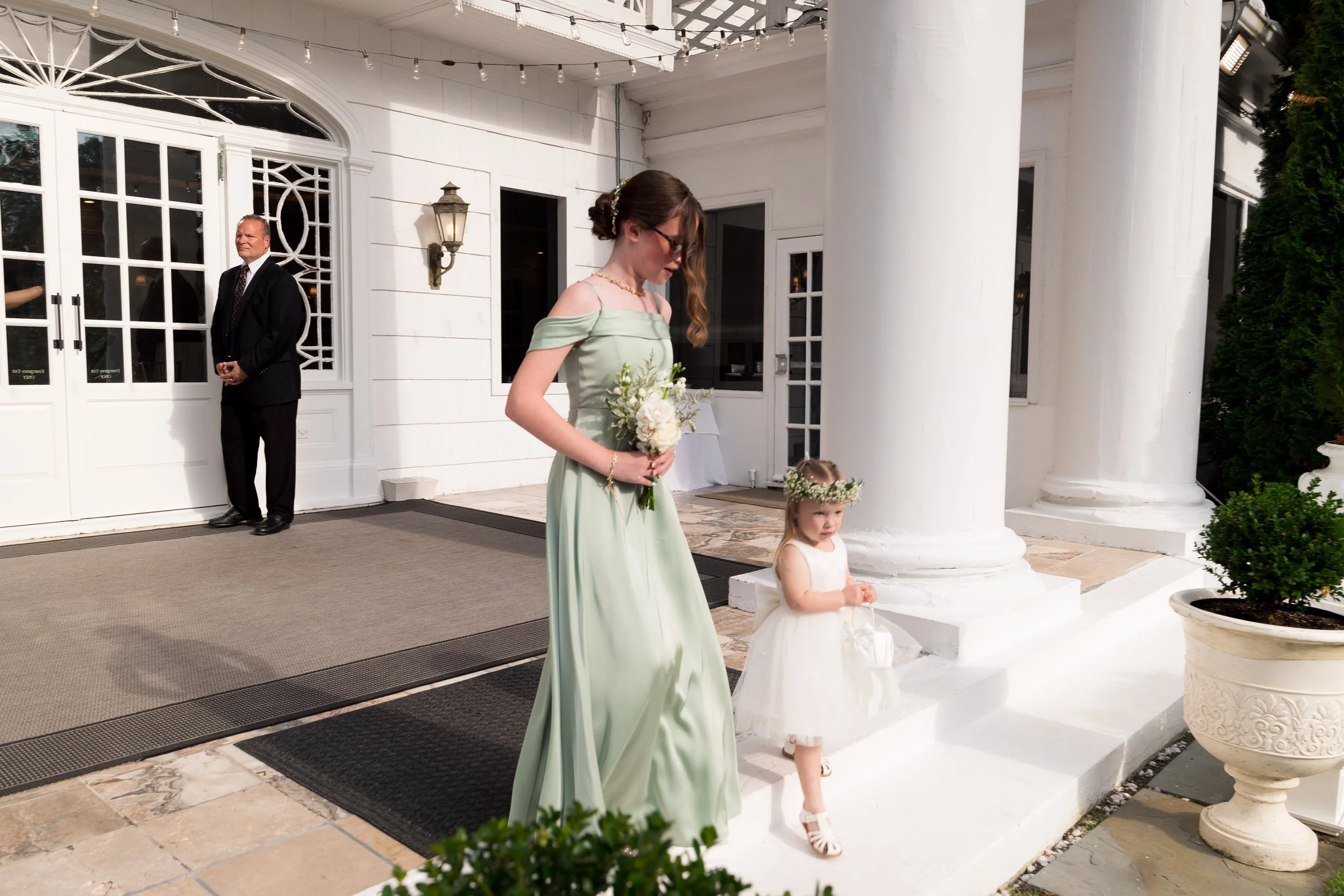 Young woman in a light green dress holding a bouquet of flowers, standing on wedding steps with a young girl in a white dress and floral headband, next to large white columns at a wedding venue, with an older man in a suit in the background.