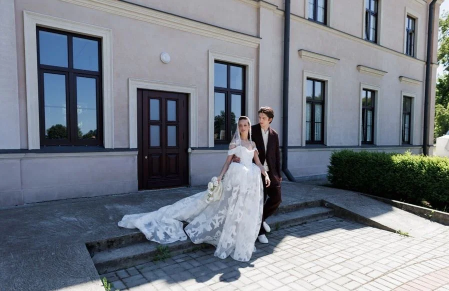 A bride in a white wedding dress and veil holding a bouquet, standing next to a groom in a dark suit, outside a beige building with multiple windows and a door, on a paved area with steps.