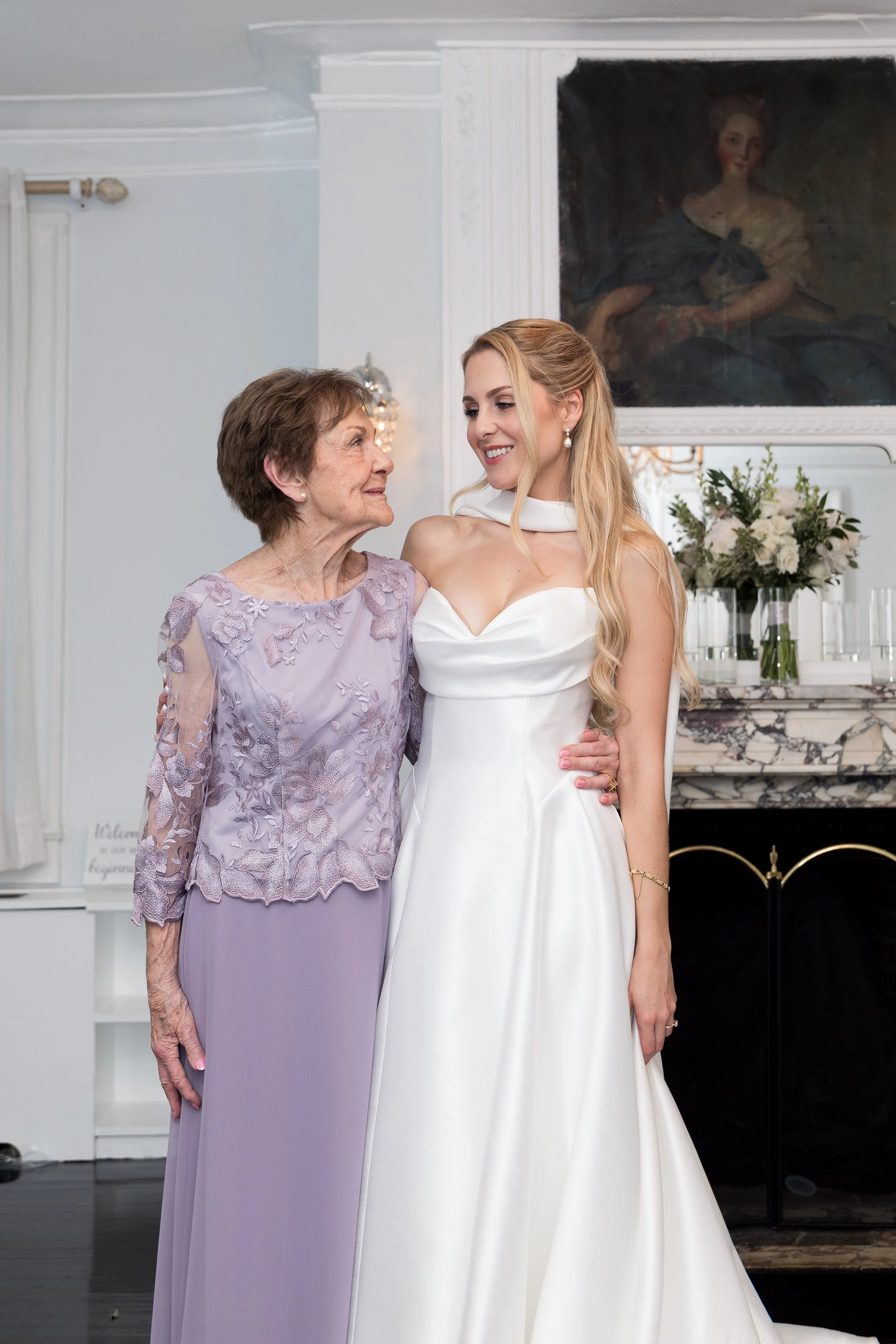 An elderly woman in a lavender dress with lace details and a young woman in a white wedding dress standing close together, smiling at each other in a bright, elegant room with a floral arrangement and a painting of a woman in historical clothing hang