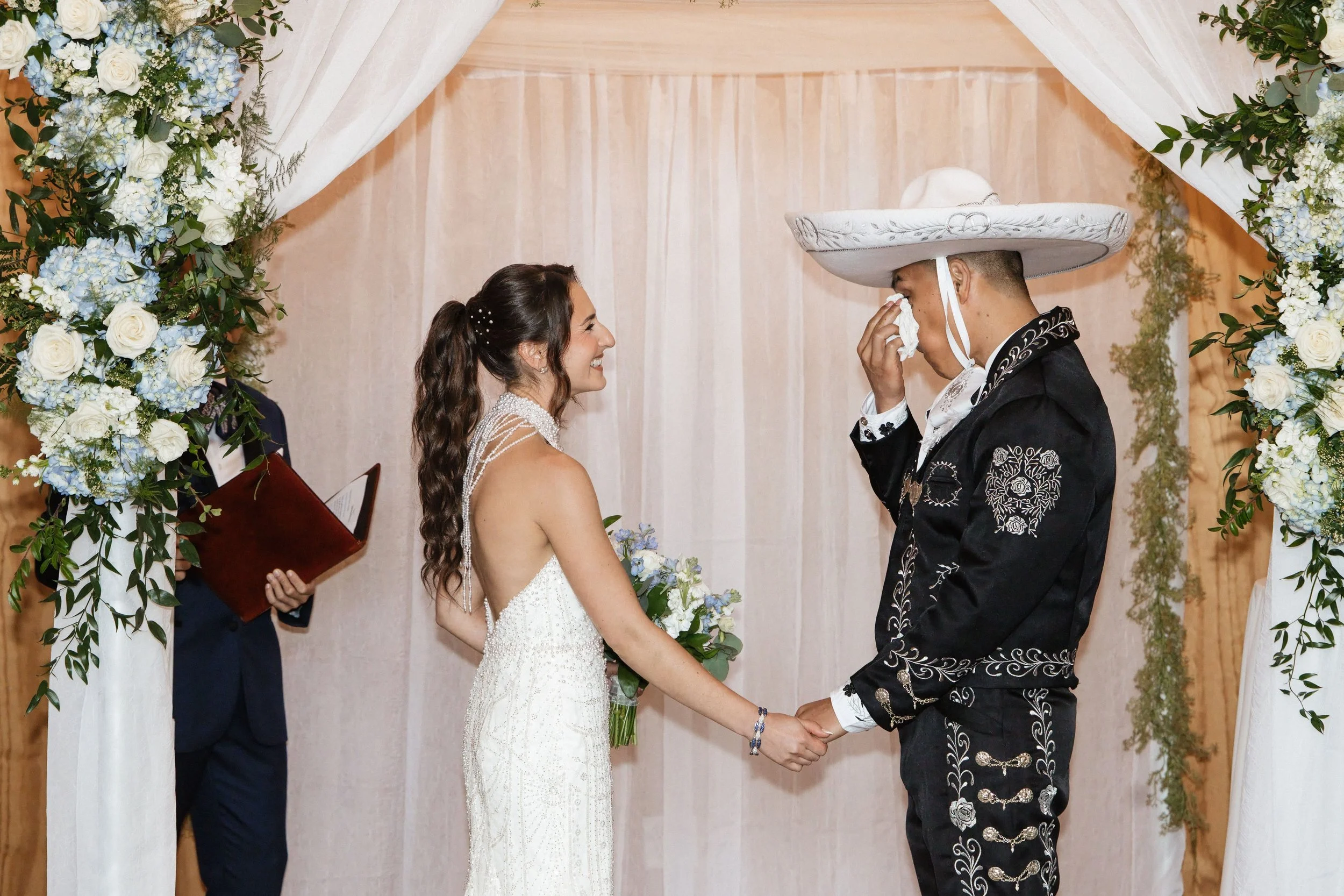 A bride and groom exchanging vows during a wedding ceremony under floral archway with white and blue flowers.
