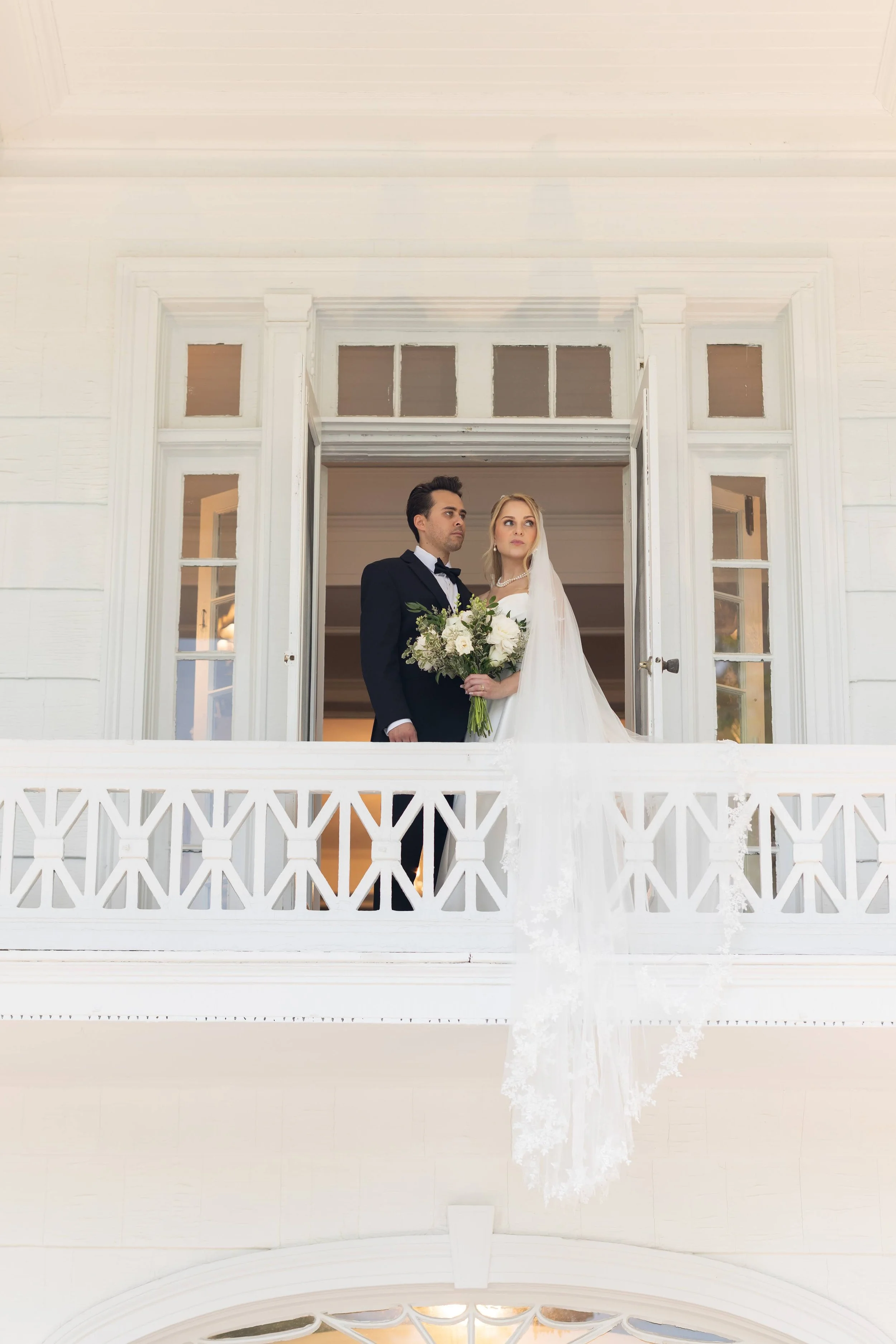 A bride and groom standing on a white porch with detailed railing, inside a bright yellow house, looking thoughtfully into the distance.