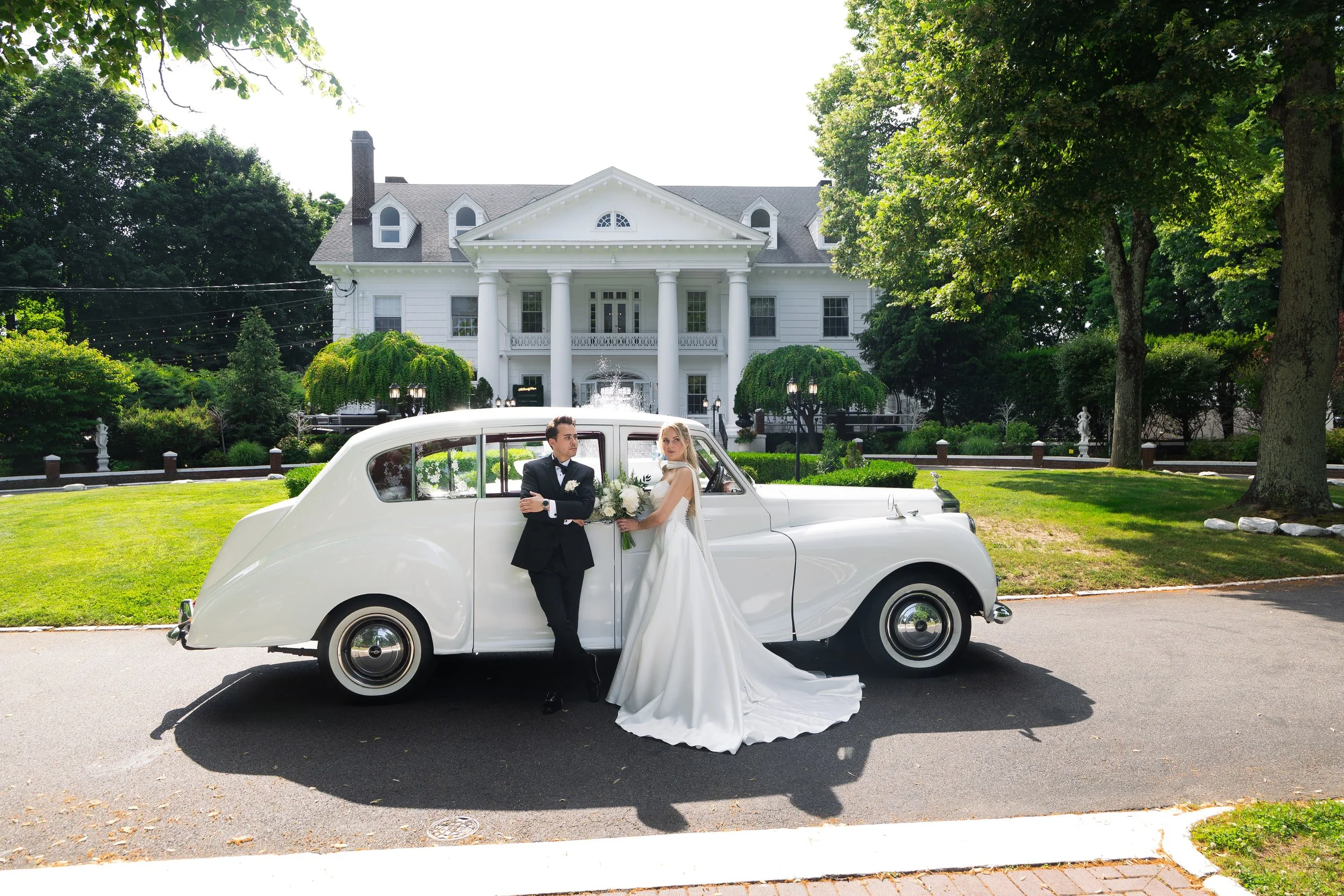 A newlywed couple posing next to a vintage white car in front of a large white mansion, surrounded by green trees and manicured bushes.