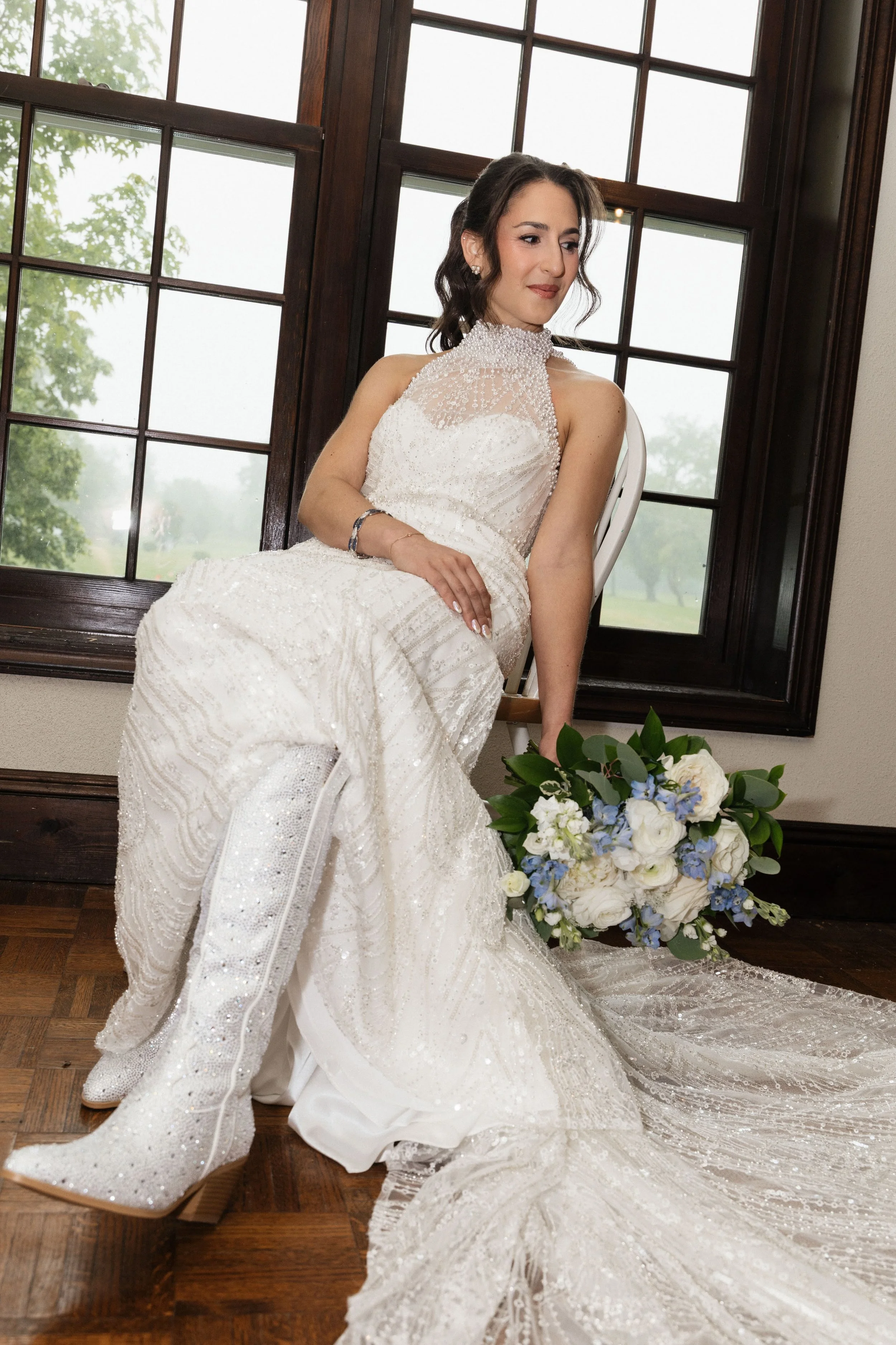 A woman in a white wedding dress sitting indoors near a large window with wooden frames, holding a bouquet of white and blue flowers on the floor.