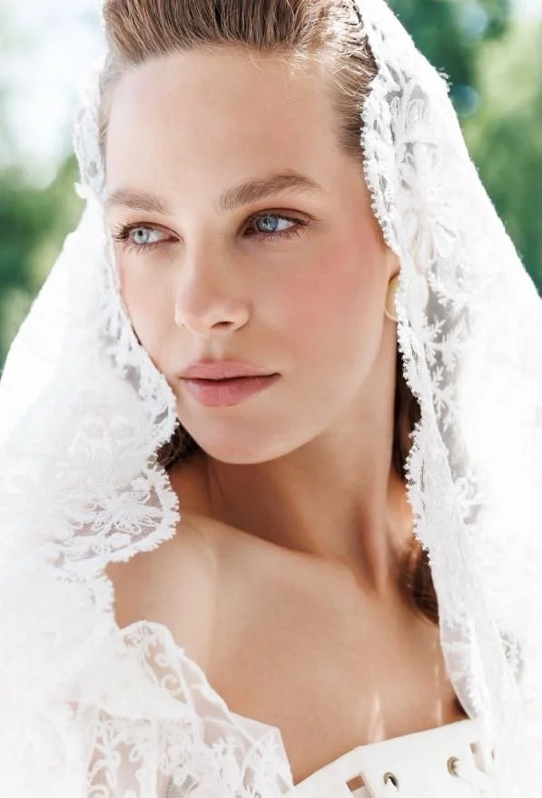 A woman with blue eyes and light hair, wearing a white lace garment and a lace veil, outdoors with blurred greenery in the background.
