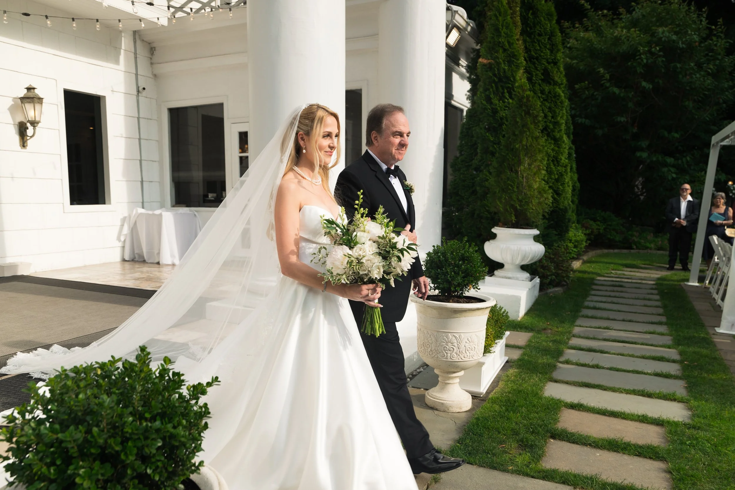 A bride in a white wedding dress holding a bouquet, walking with an older man in a black tuxedo outdoors during a wedding ceremony. There are guests in the background and greenery around.