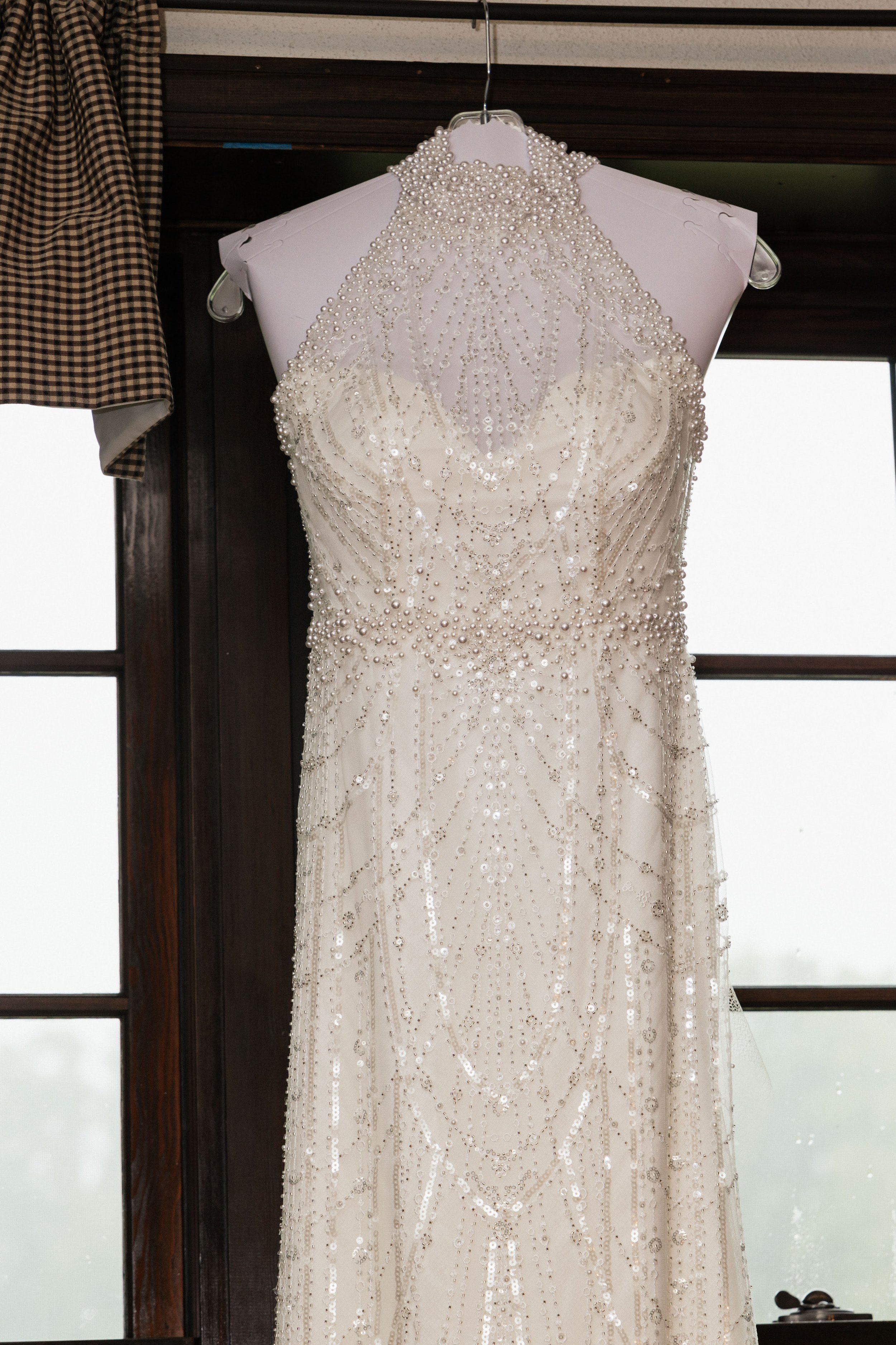 A white wedding dress with pearl and sequin embellishments hanging on a white hanger in front of dark wood and window frames.
