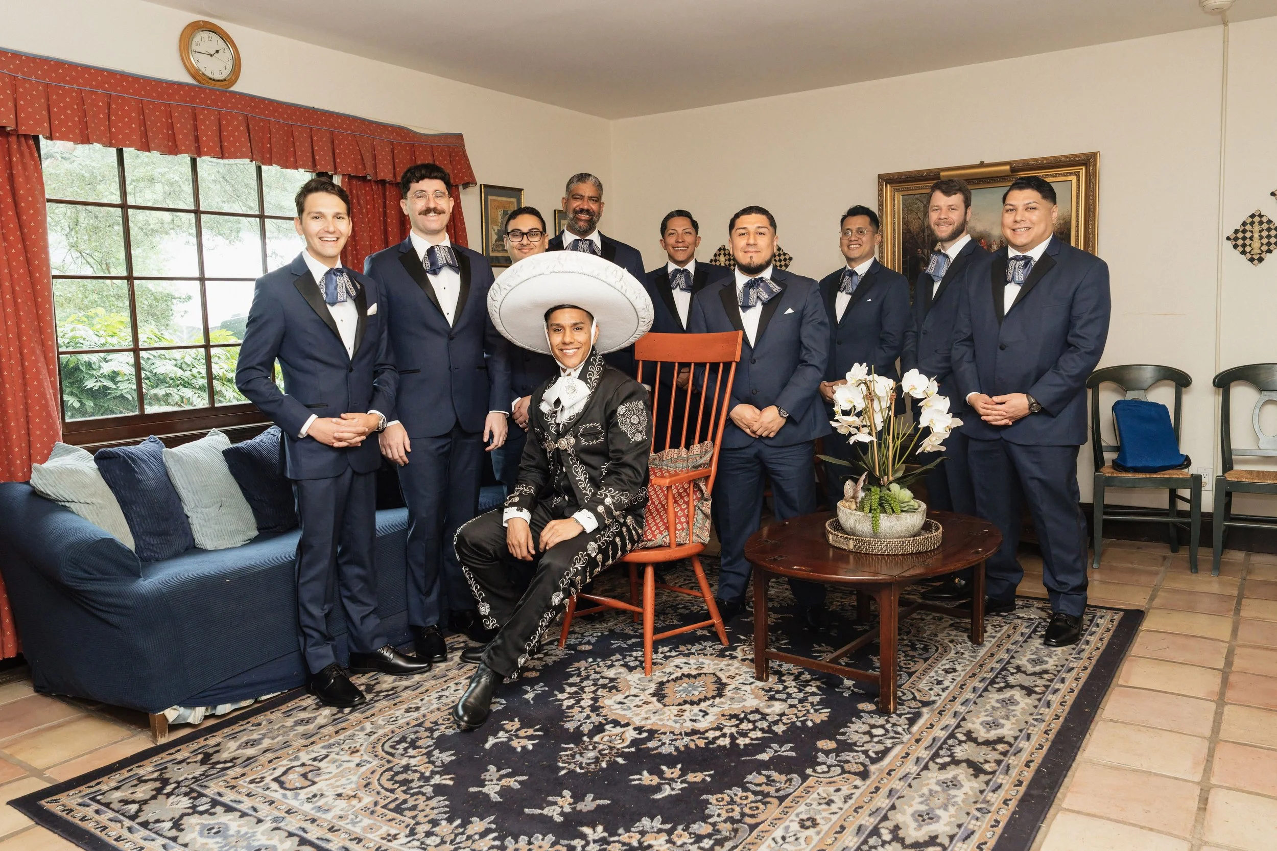 A group of ten men dressed in dark blue suits and bow ties, with one seated wearing traditional Mexican attire and a large sombrero. They are inside a living room with a window, curtains, a patterned rug, and artwork on the walls, all smiling for the