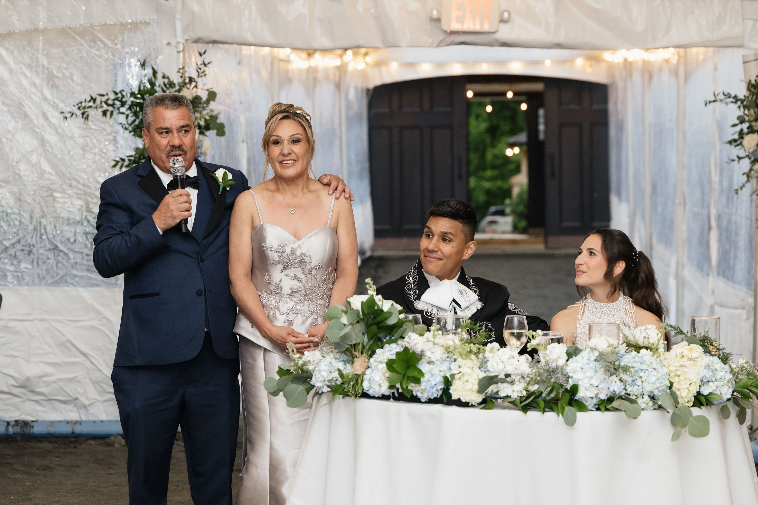 A man in a navy suit giving a speech at a wedding reception, with a woman in a wedding dress standing next to him, seated guests, wedding decorations, and floral arrangements on the table.