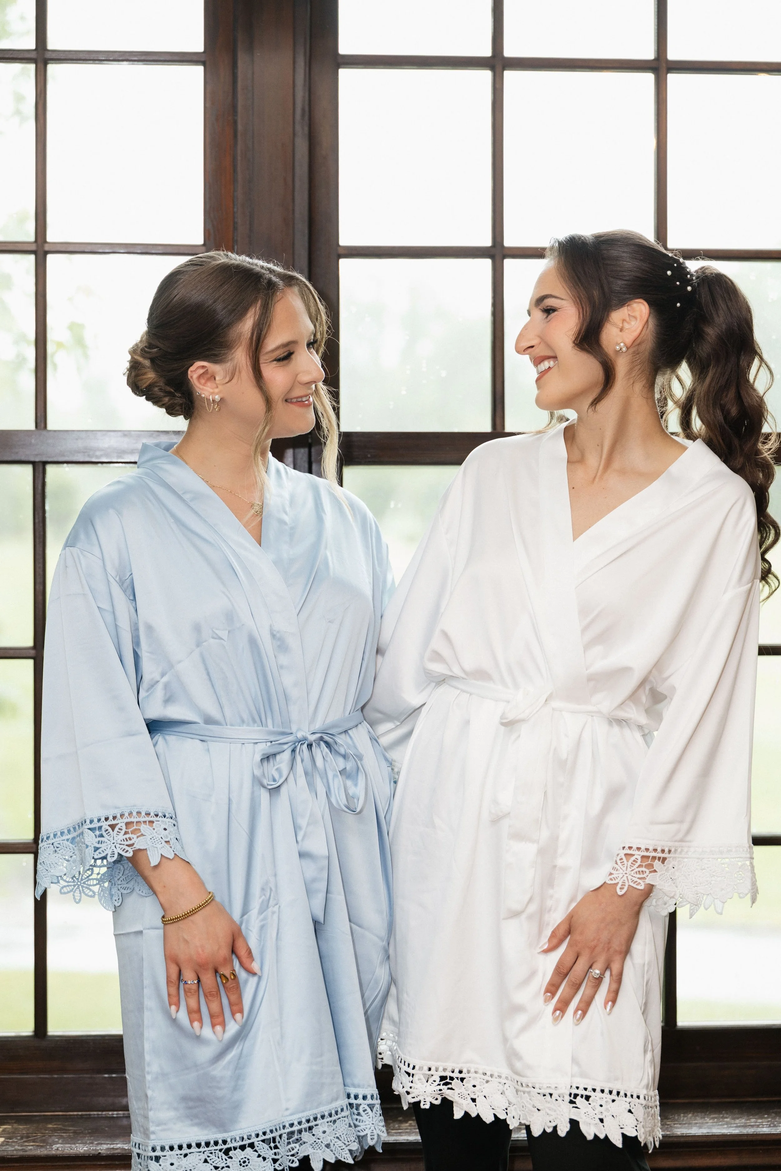 Two women standing indoors in front of large windows, smiling and looking at each other. Both are wearing light-colored robes with lace trim, one in blue, the other in white.