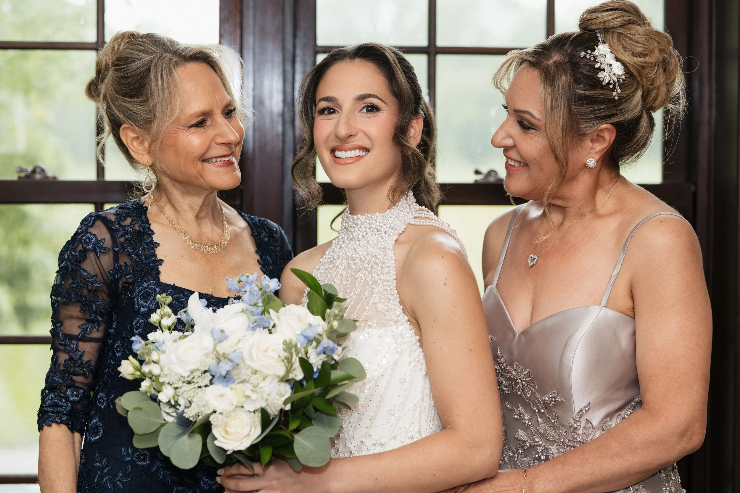 A bride in a white wedding dress holding a bouquet of white and light blue flowers, flanked by two women in elegant dresses, smiling in front of large windows.
