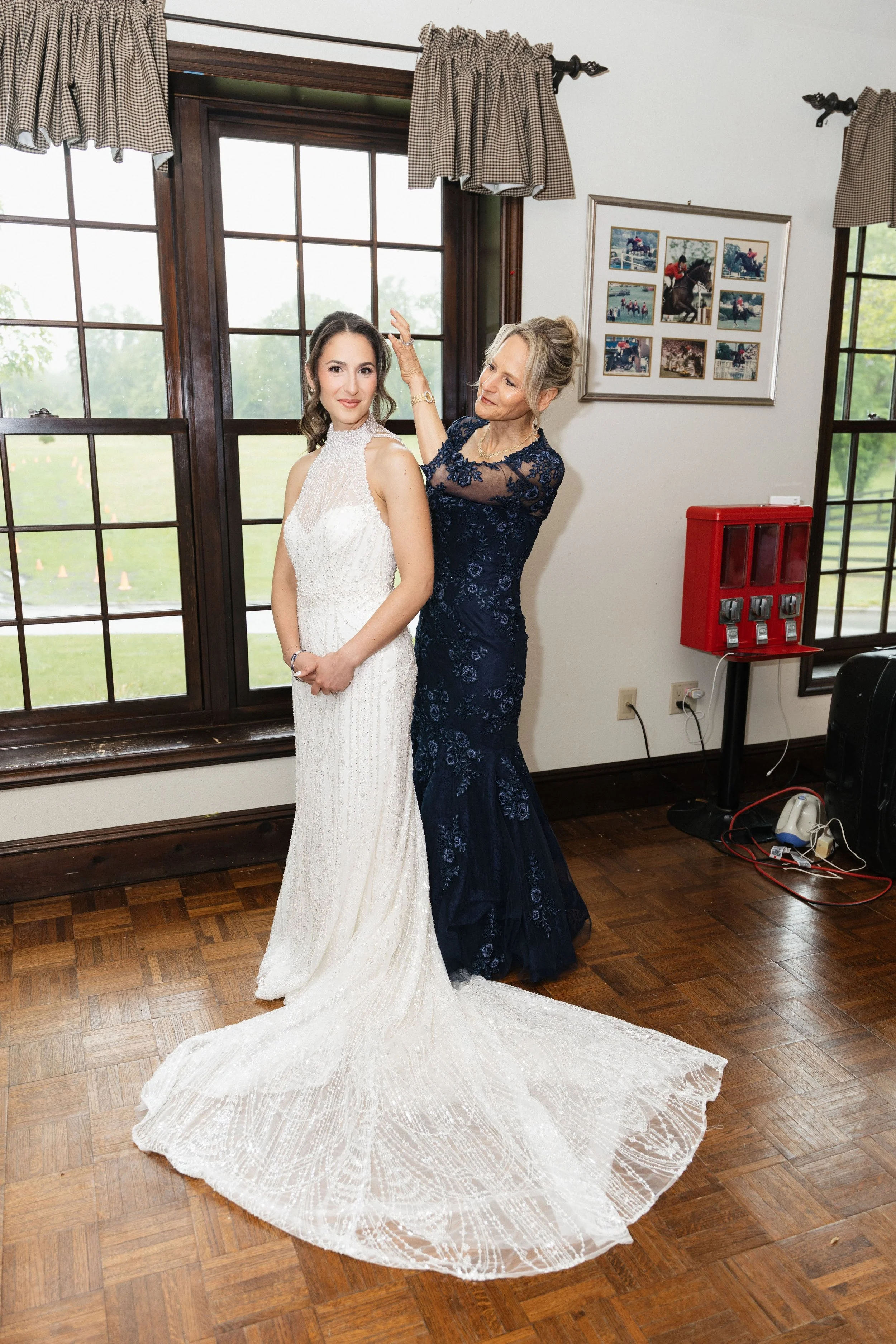 A bride in a white gown with lace details stands next to an older woman in a navy blue dress with floral embroidery, who is adjusting the bride's hair. They are inside a room with large windows, wooden floors, and a wall decorated with a framed colla