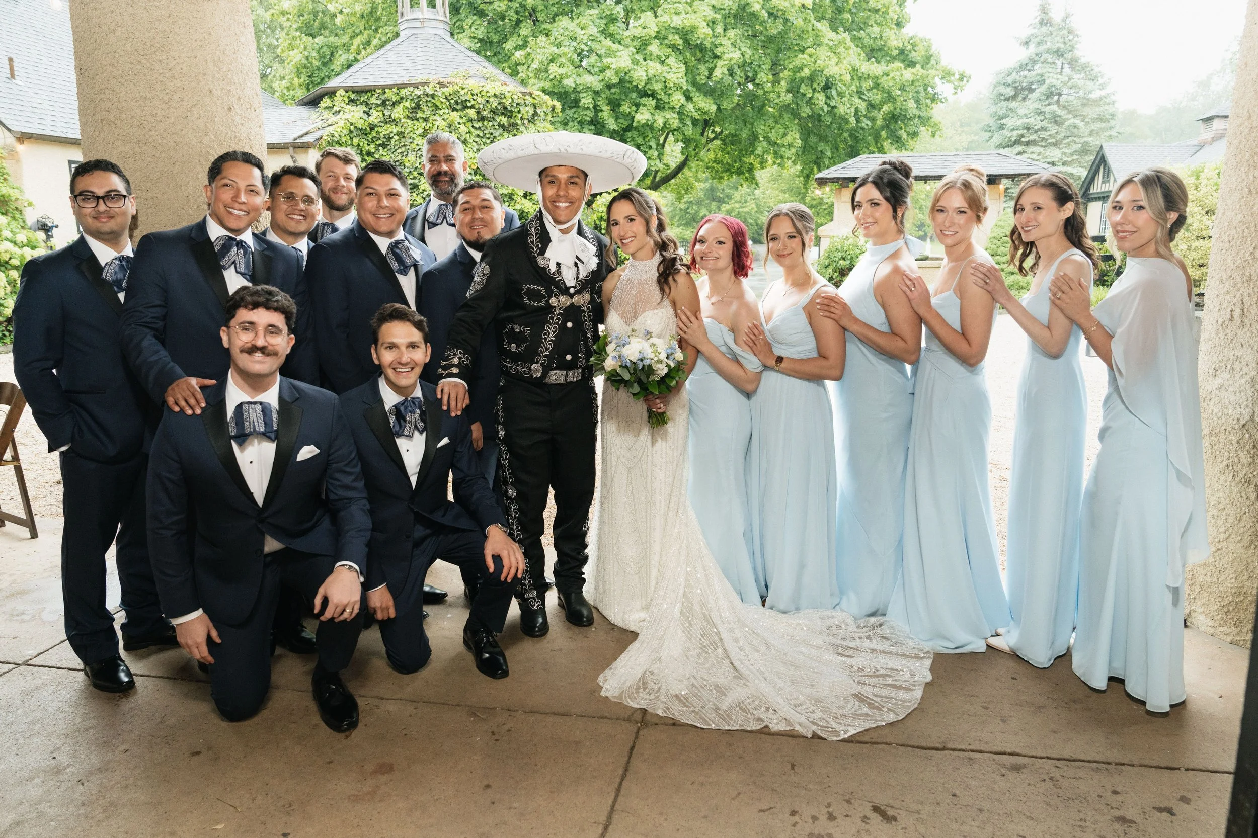 A wedding group of men and women posing outdoors on a porch, with the bride in a white dress holding a bouquet and the groom in a charro suit, surrounded by bridesmaids in light blue dresses and groomsmen in dark suits, with trees and houses in the b