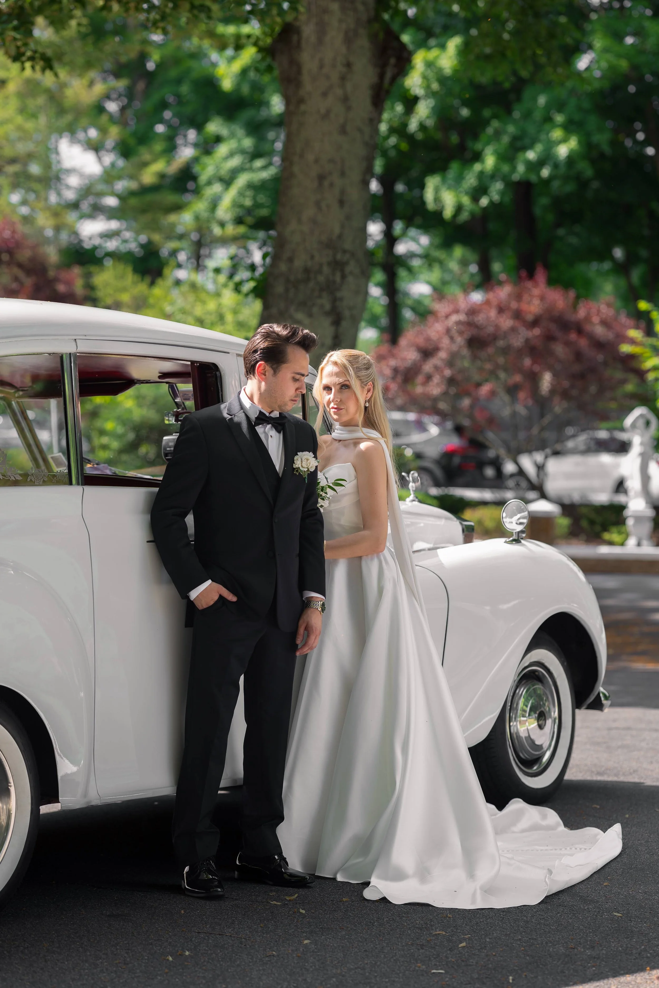 A man and woman dressed in formal wedding attire leaning against a vintage white car in a park with green trees.