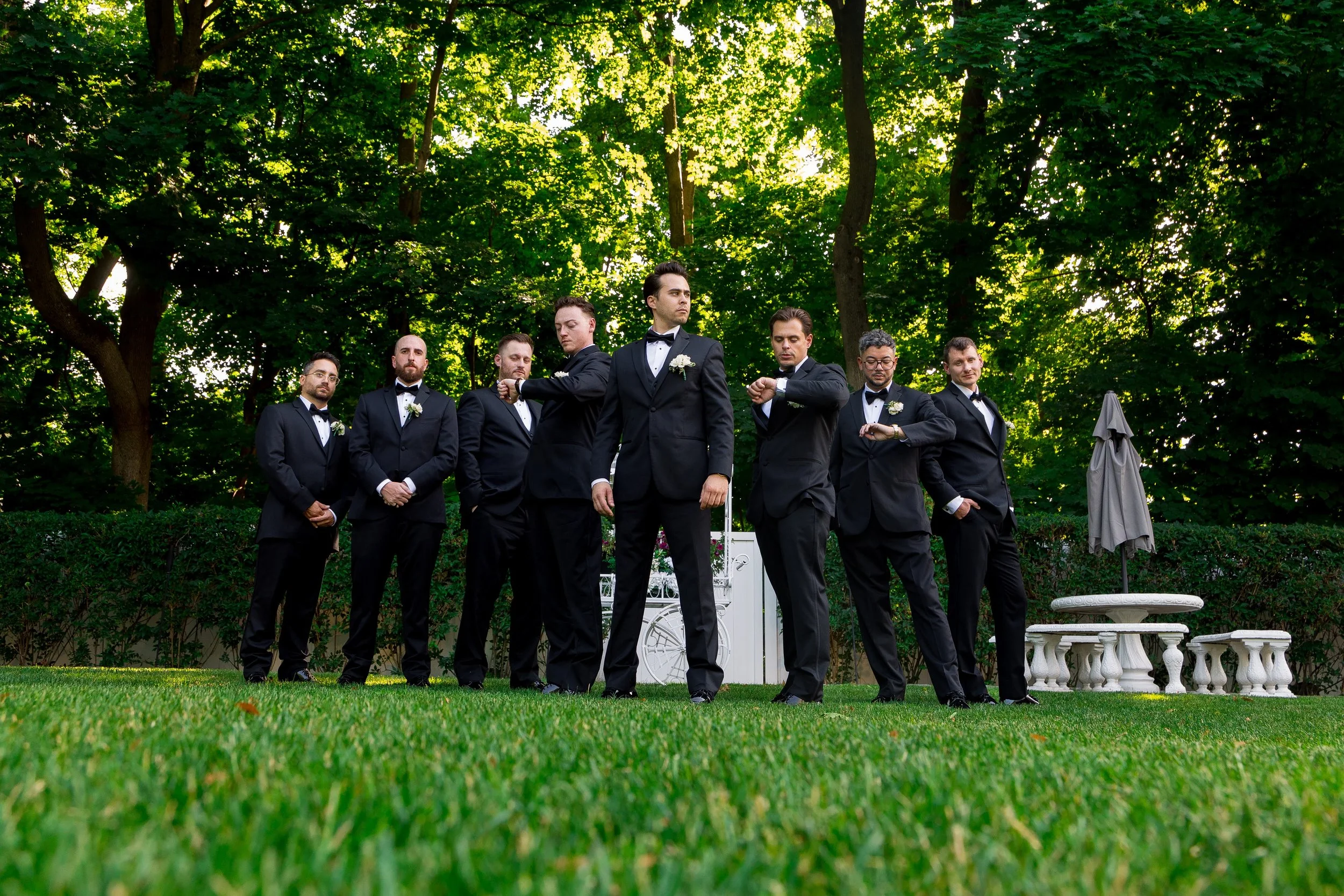 A group of eight men dressed in black tuxedos with bow ties standing outdoors in a row in front of a lush green canopy of trees, with some checking their watches and others looking ahead.