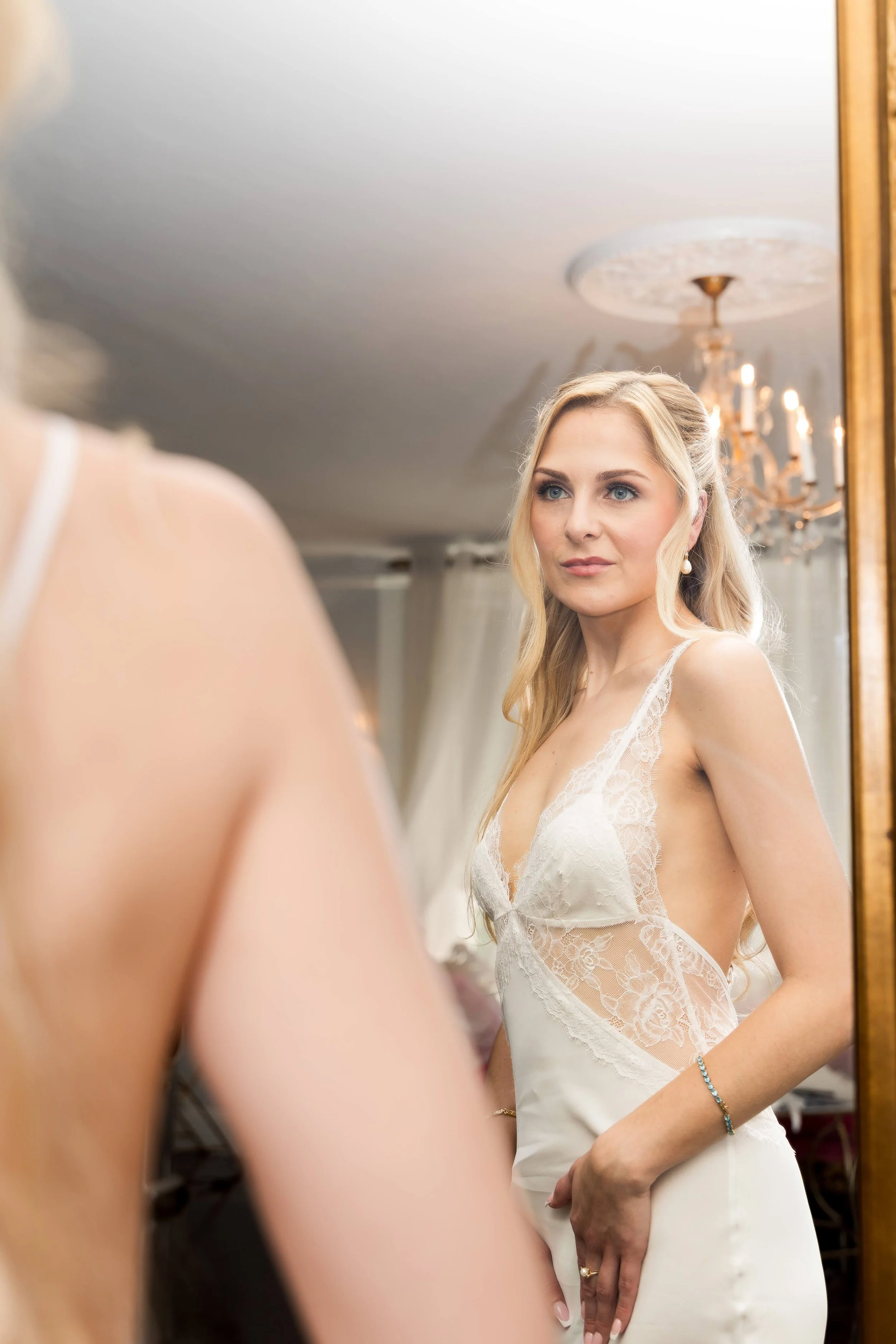 A woman in a wedding dress looking in a mirror, with a chandelier and other wedding preparations in the background.