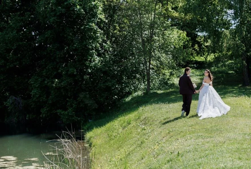 Couple dressed in wedding attire holding hands in a grassy park near a small pond with trees in the background.