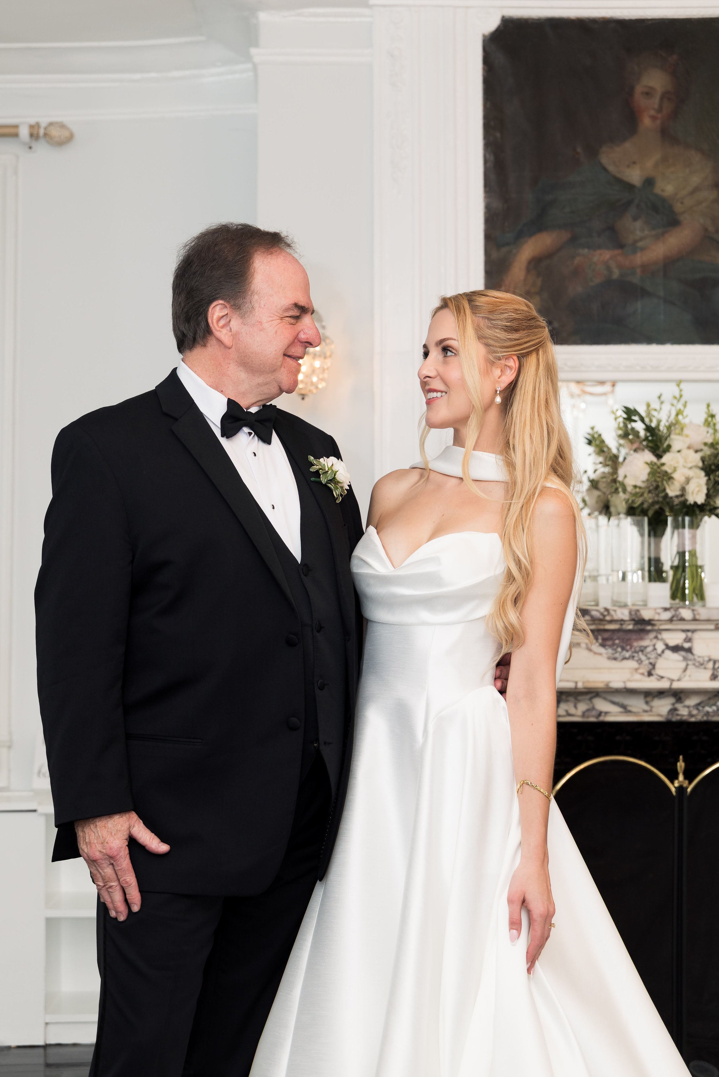 A man in a black tuxedo and a woman in a white gown looking at each other, smiling, in a decorated room with floral arrangements and a painting in the background.