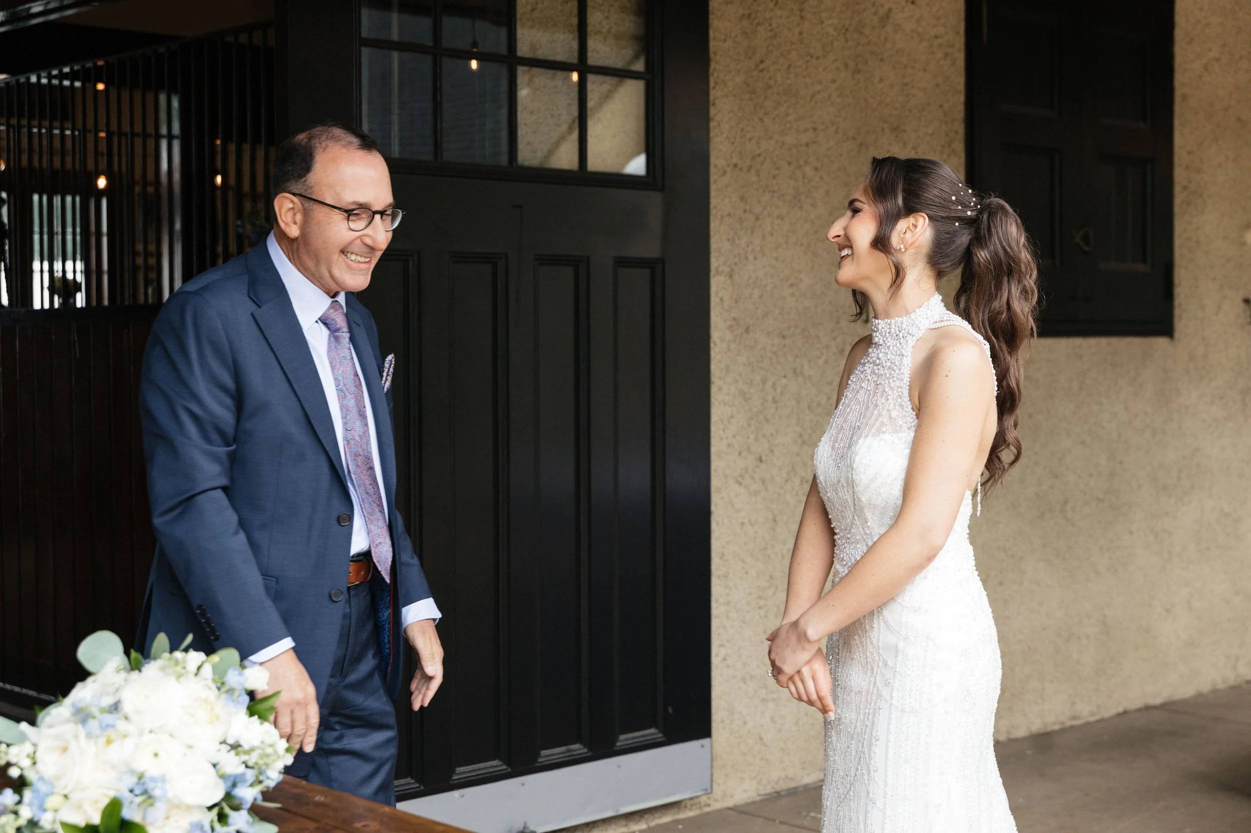 A bride and an older man are smiling and facing each other outside a building during a wedding celebration.