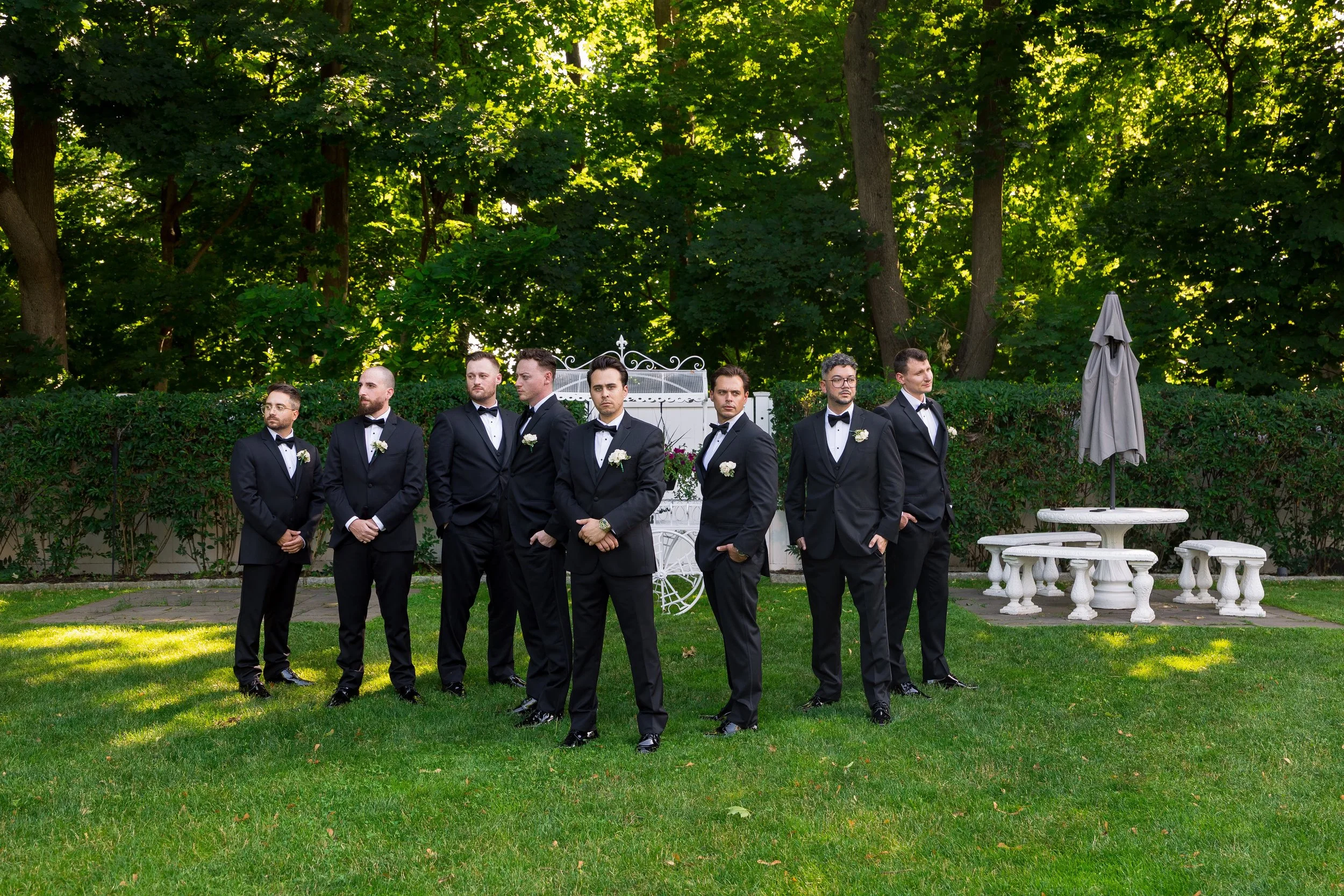 A group of seven men dressed in formal black tuxedos with bow ties and white boutonnieres standing outdoors on a well-kept lawn, with lush green trees in the background and a white wrought-iron bench and table with an umbrella on the right side.