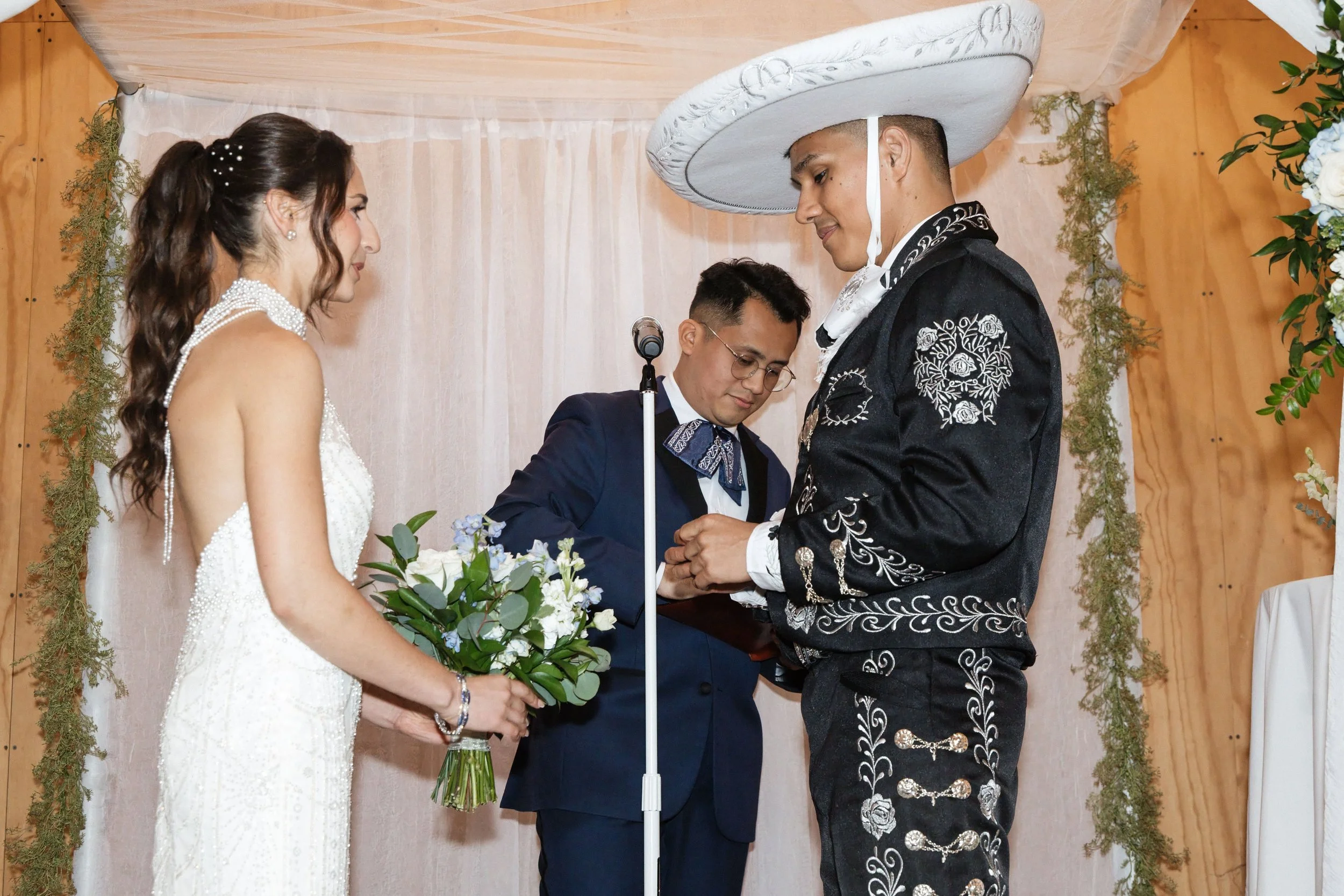 A couple getting married with a woman officiating. The groom is wearing a traditional Mexican charro suit with silver embroidery and a large sombrero, and the bride is wearing a white wedding dress holding a bouquet of flowers.