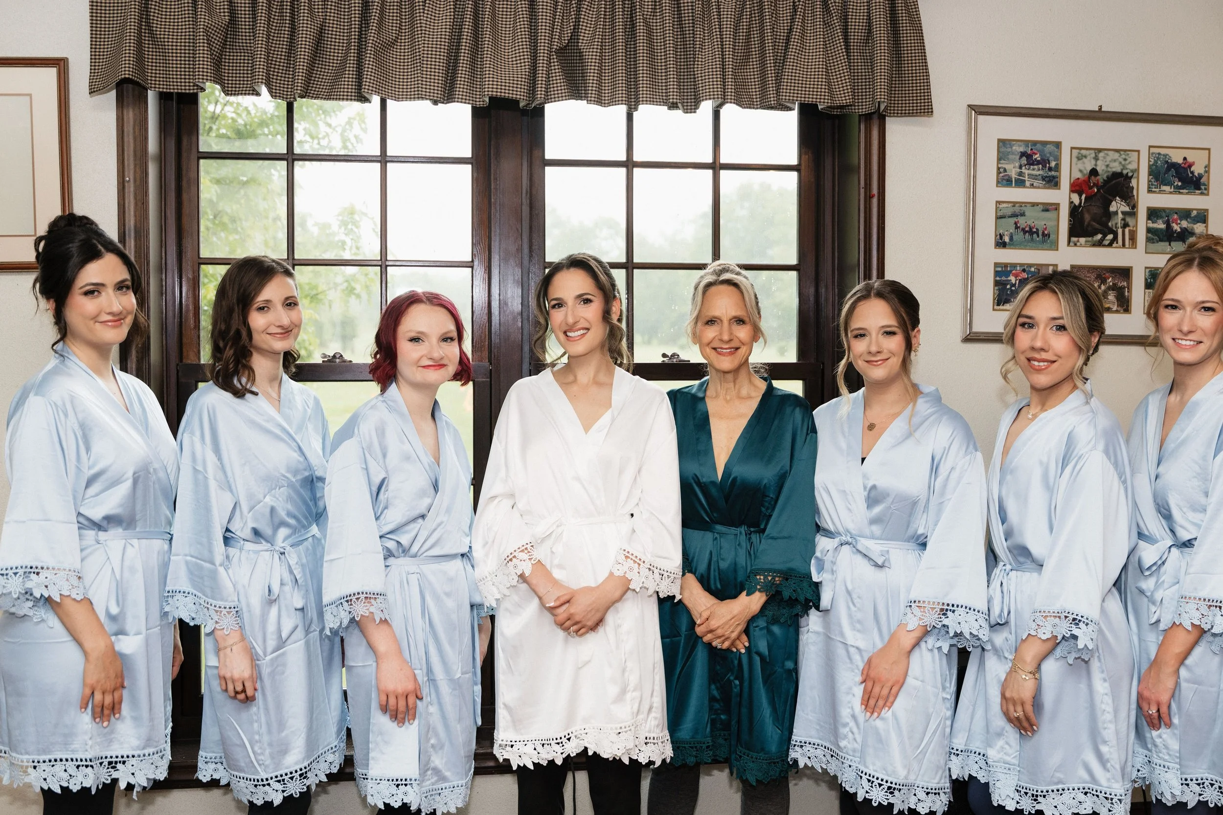 Group of women standing inside near a large window, some wearing matching light blue robes, others in different attire.