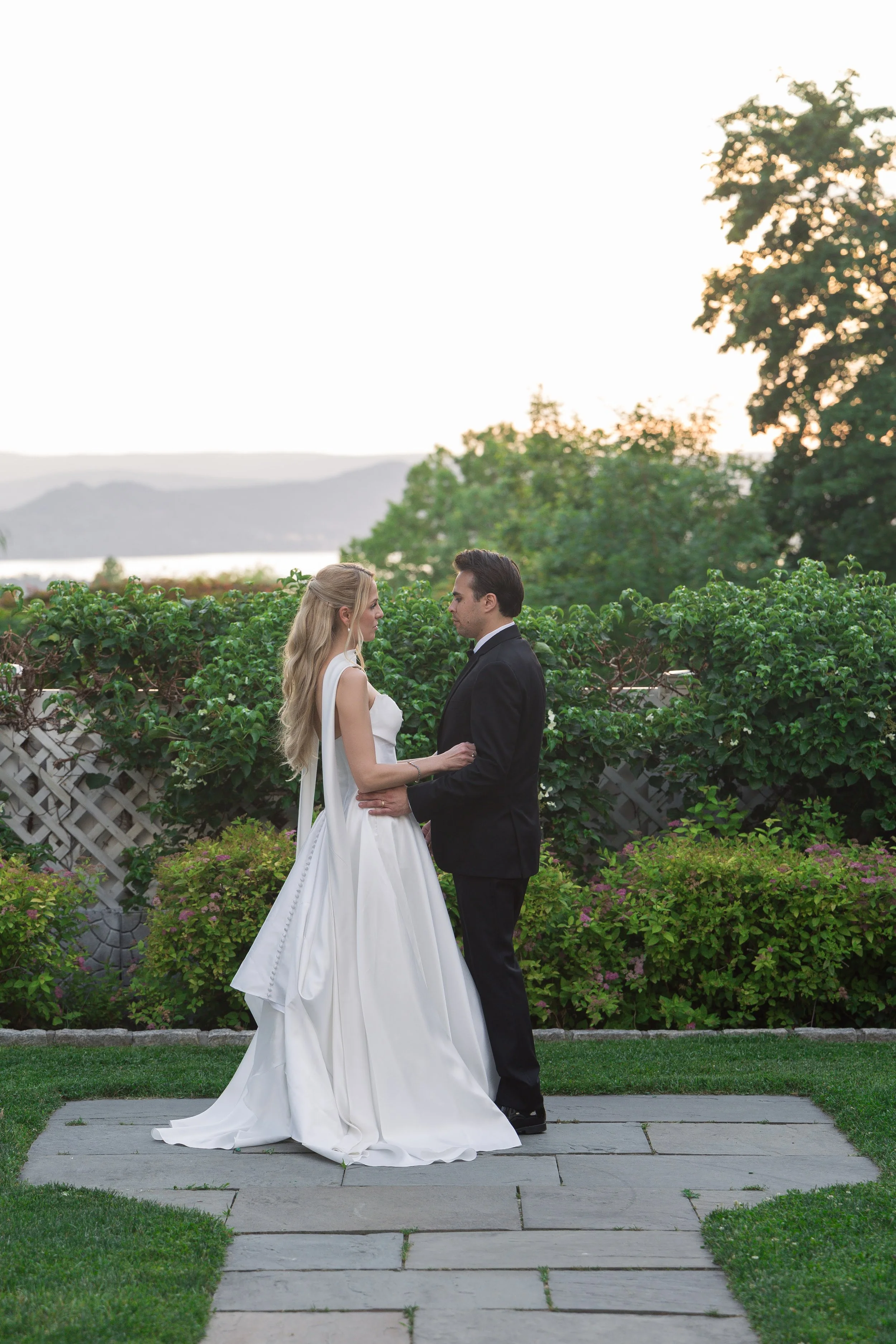 A bride and groom standing close together outdoors, holding hands, with a scenic background of trees, hills, and a body of water at sunset.