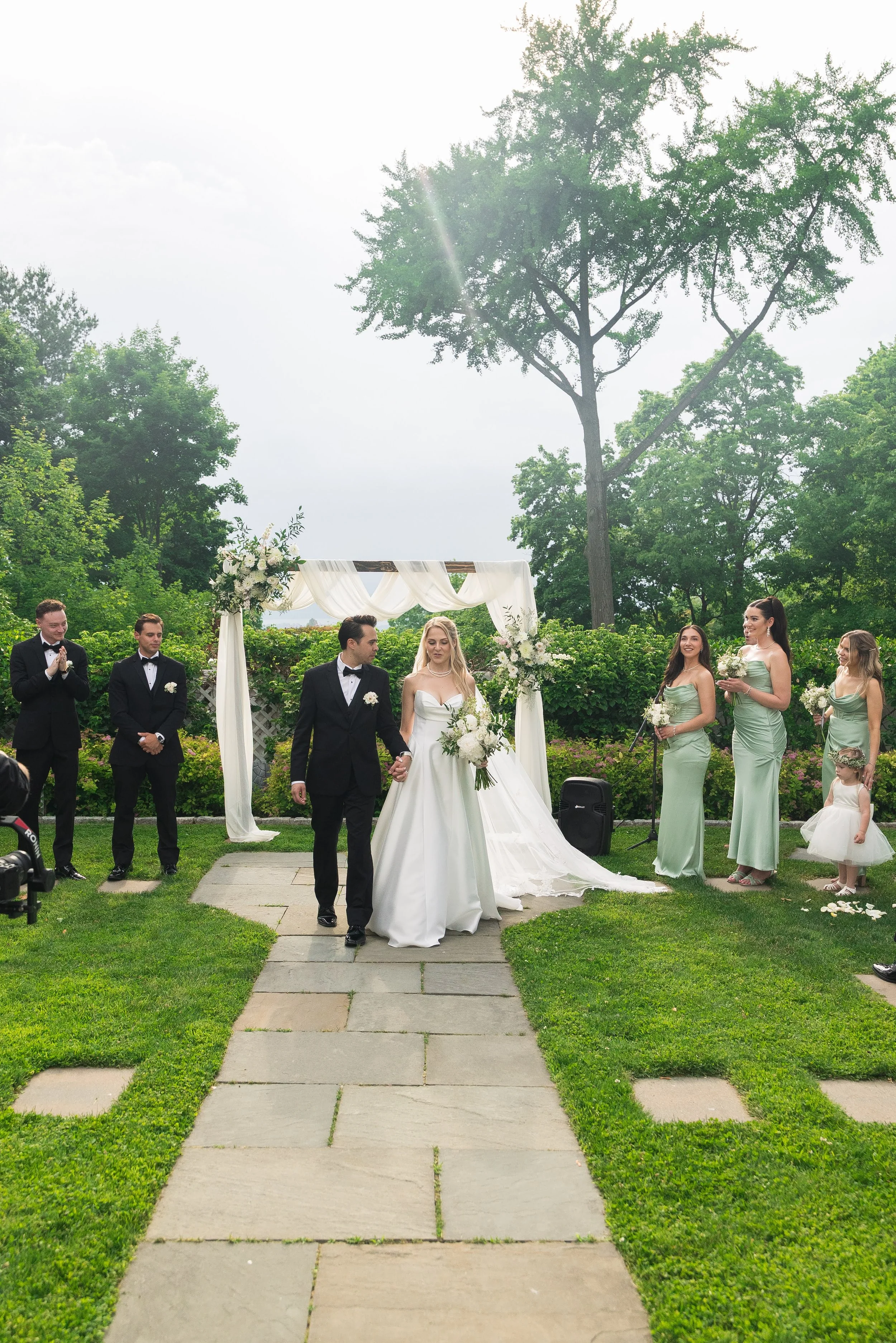 A wedding ceremony outdoors in a garden with a green lawn and tall trees. The bride and groom stand under a white arch with draped fabric, surrounded by bridesmaids in matching light green dresses and groomsmen in black tuxedos. The scene is illumina
