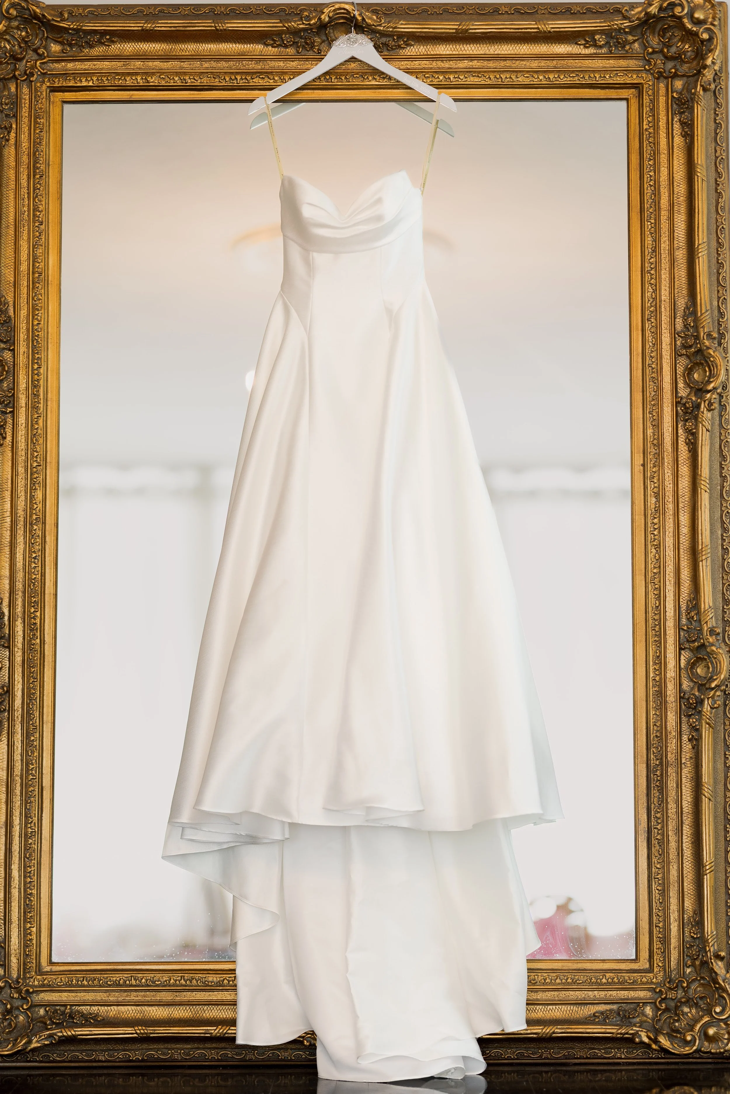 A white wedding dress hanging on a white hanger inside an ornate gold picture frame.