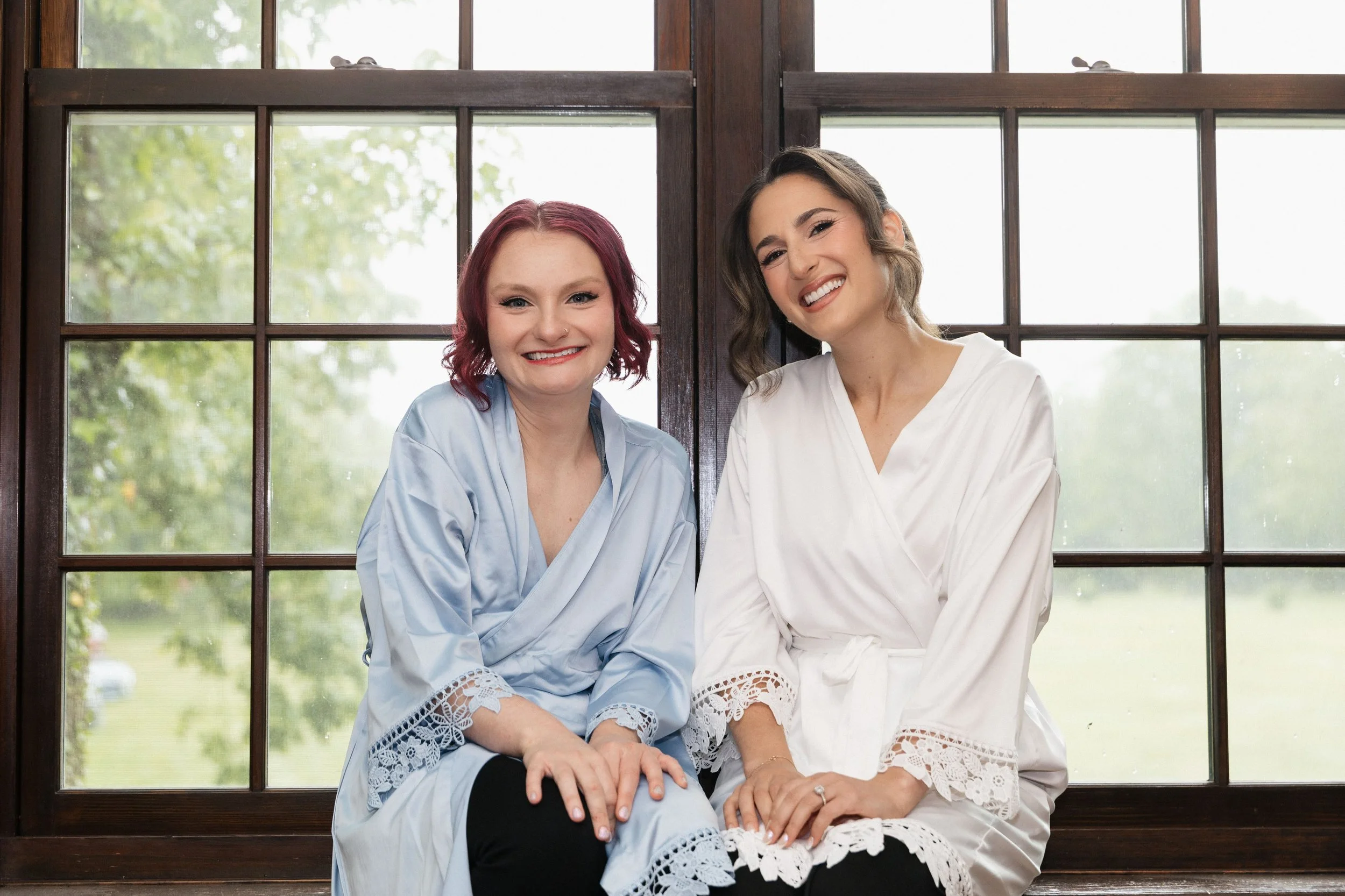 Two smiling women sitting together by a window with wooden frames, dressed in light-colored clothing, with a green outdoor scene visible outside.