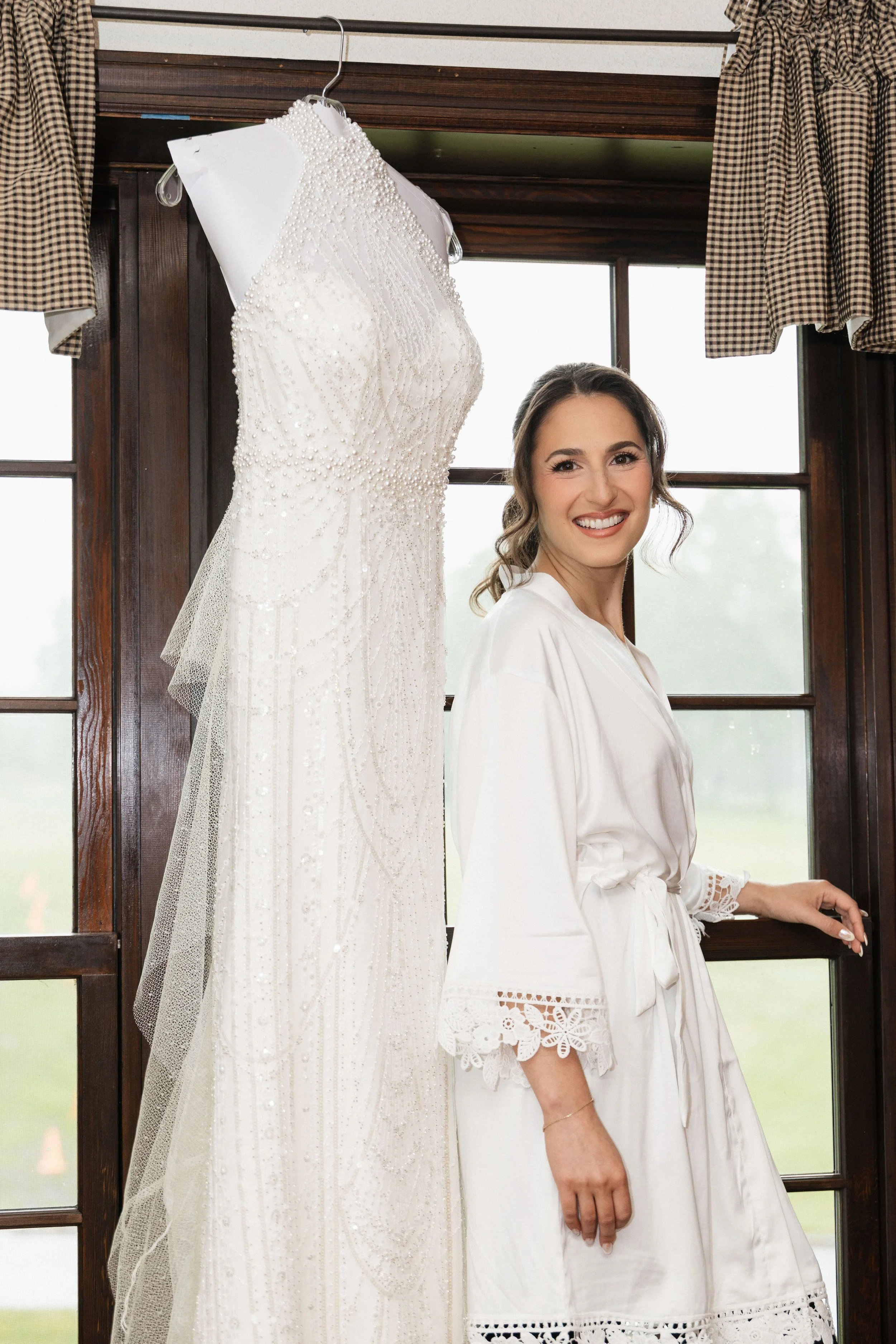 A woman in a white robe smiling while standing near a window, with a white wedding dress hanging next to her.
