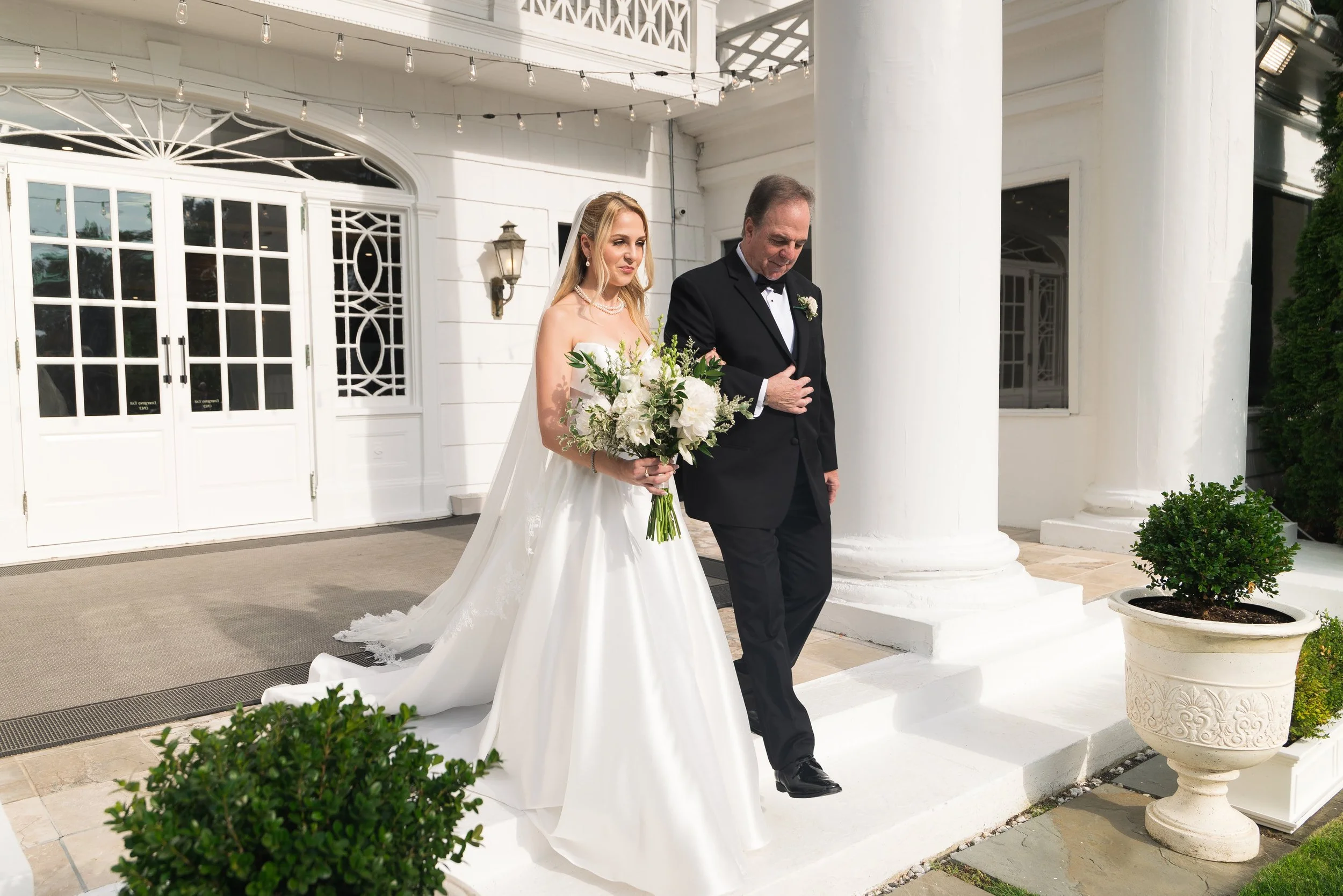 A bride in a white wedding dress holding a bouquet of white flowers walking down the steps with an older man in a black tuxedo, outside a white building with large columns and potted plants.