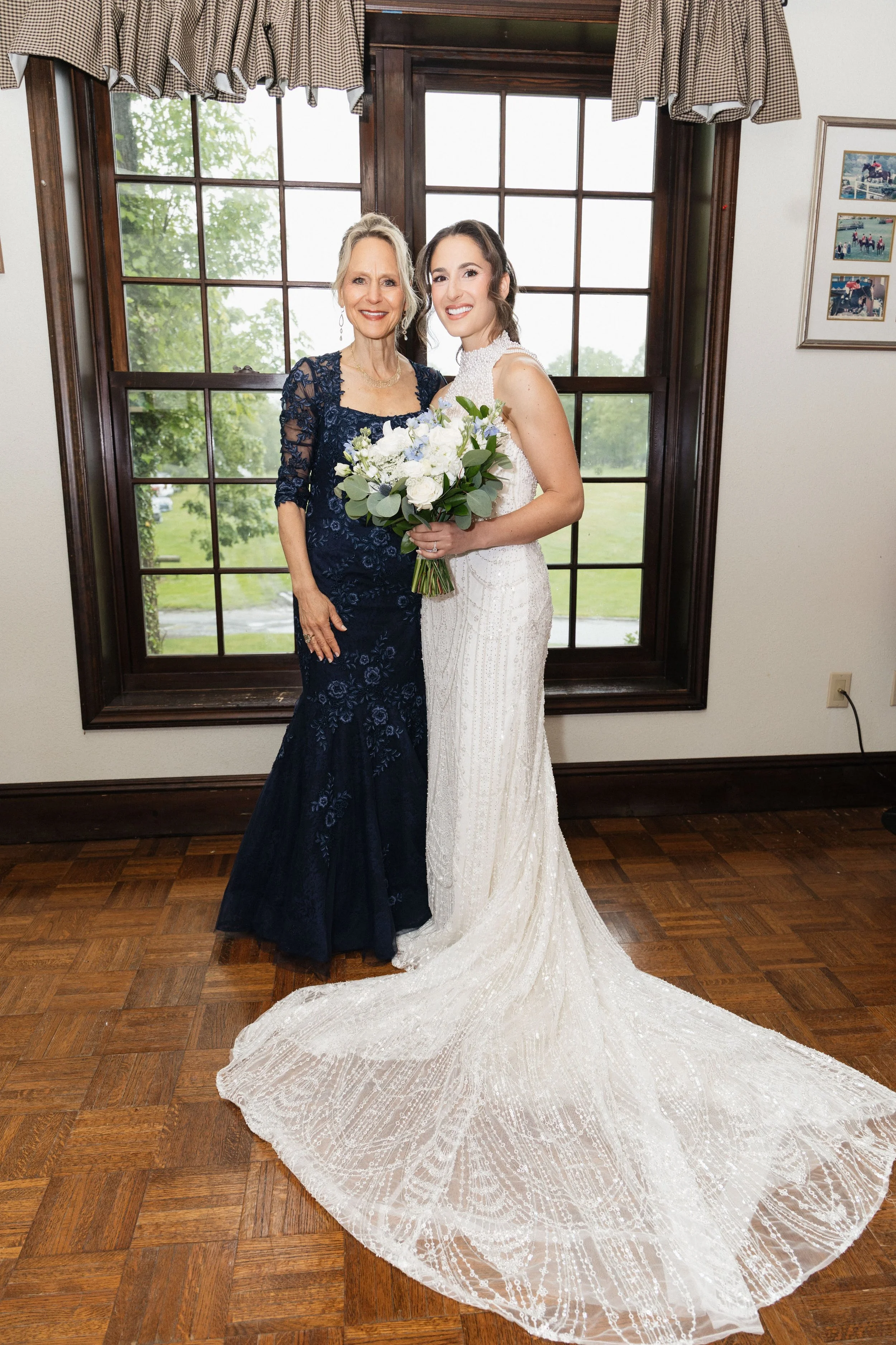 A bride in a white wedding gown holding a bouquet of white and lavender flowers, standing with an older woman in a navy blue lace dress in front of a window with wood framing and checked valances, smiling.