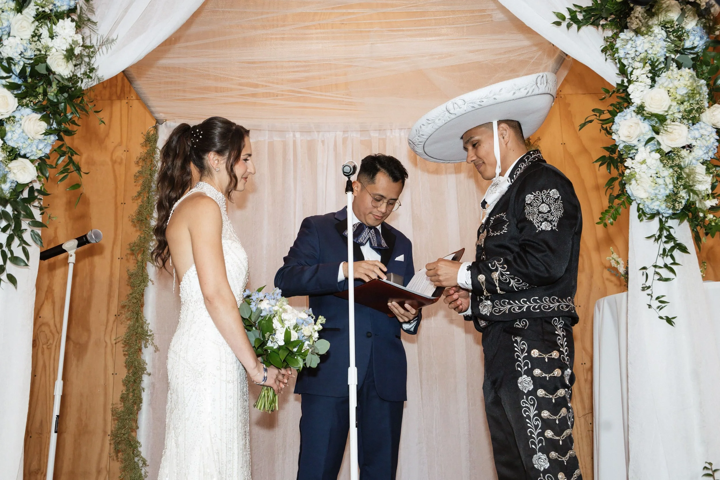 A couple getting married, holding hands, surrounded by their officiant and clergy, during a wedding ceremony with floral decorations.