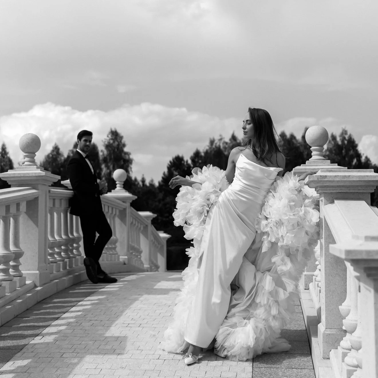 Black and white photo of a bride in a wedding gown with ruffled skirt, standing on a stone bridge railing, while a groom in a suit stands in the background leaning on the bridge railing, outdoors with trees and clouds in the sky.