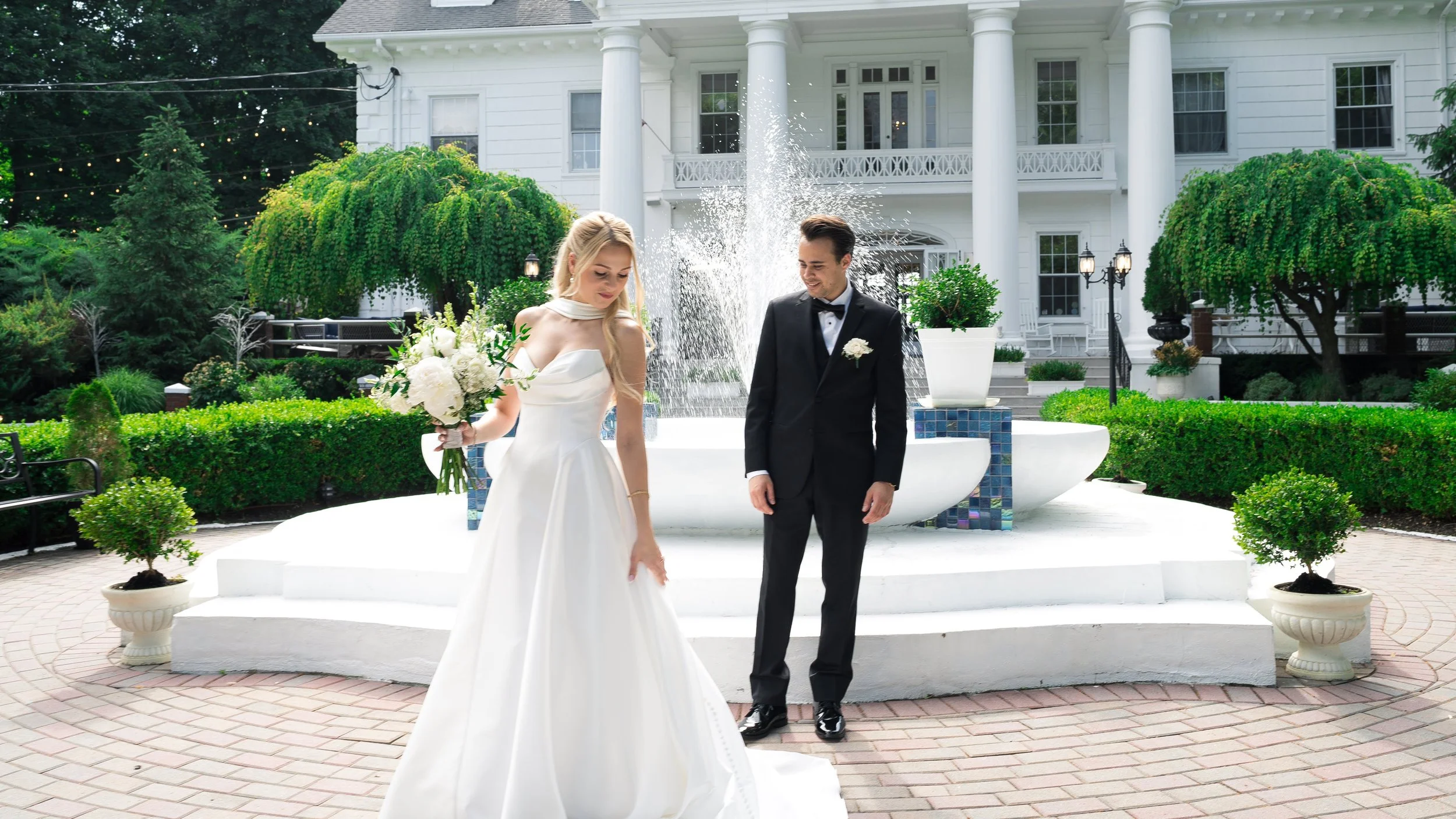 A bride and groom stand in front of a white house with green trees and bushes. The bride is wearing a white wedding dress and holding a bouquet, and the groom is dressed in a black tuxedo, both smiling.