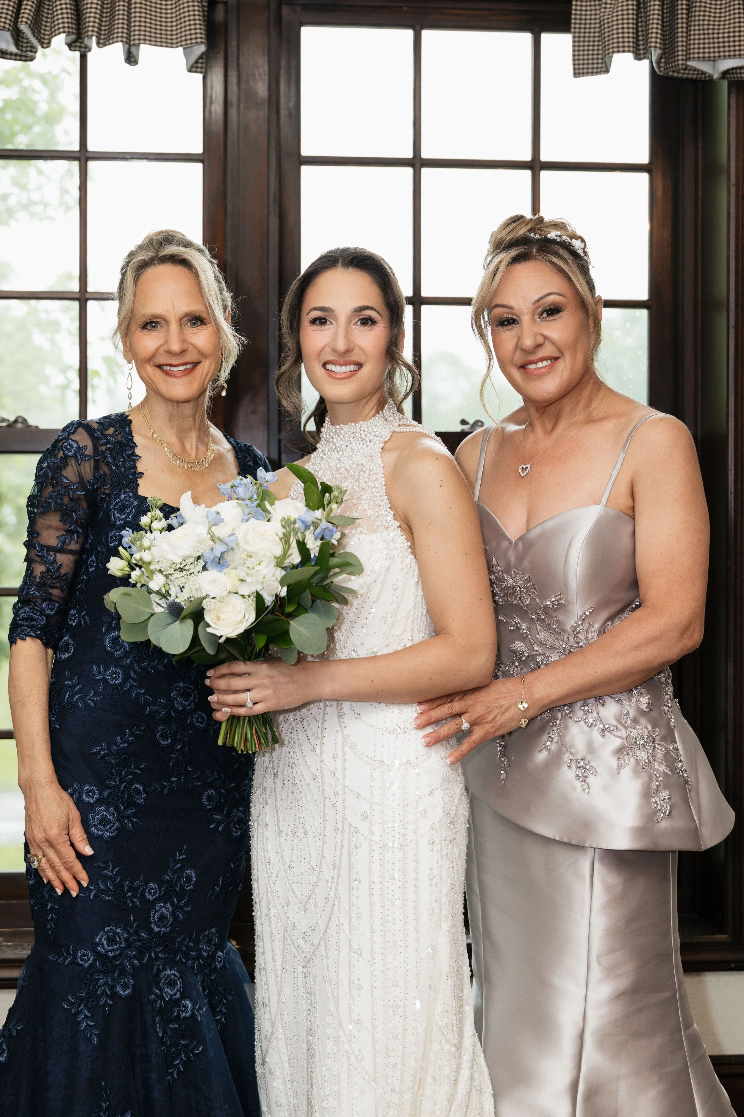 Three women standing together indoors, smiling for a photo. The woman in the center is wearing a white wedding dress and holding a bouquet of flowers. The woman on the left is wearing a dark blue dress with floral lace, and the woman on the right is 