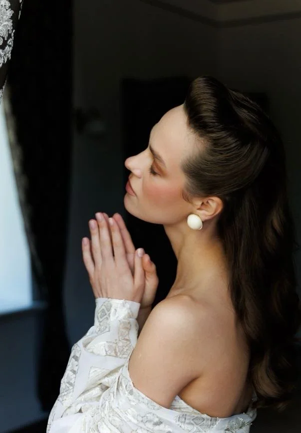 A woman with styled brown hair, wearing white earrings, and an off-shoulder lace dress, is standing inside a dimly lit room with her hands pressed together in prayer or contemplation, facing a window with curtains.