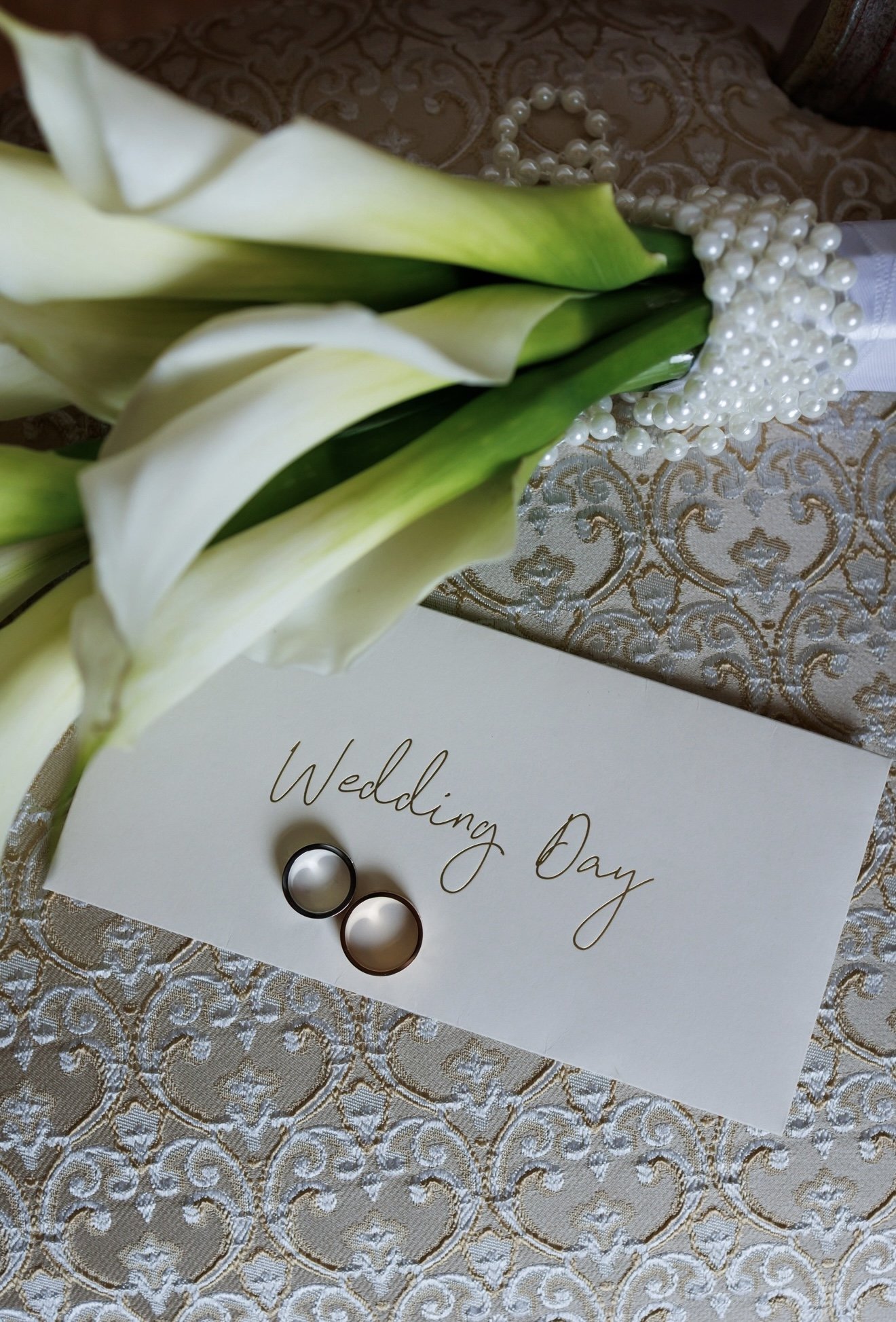 White calla lilies, wedding rings, a pearl necklace, and a wedding program on an ornate tablecloth.