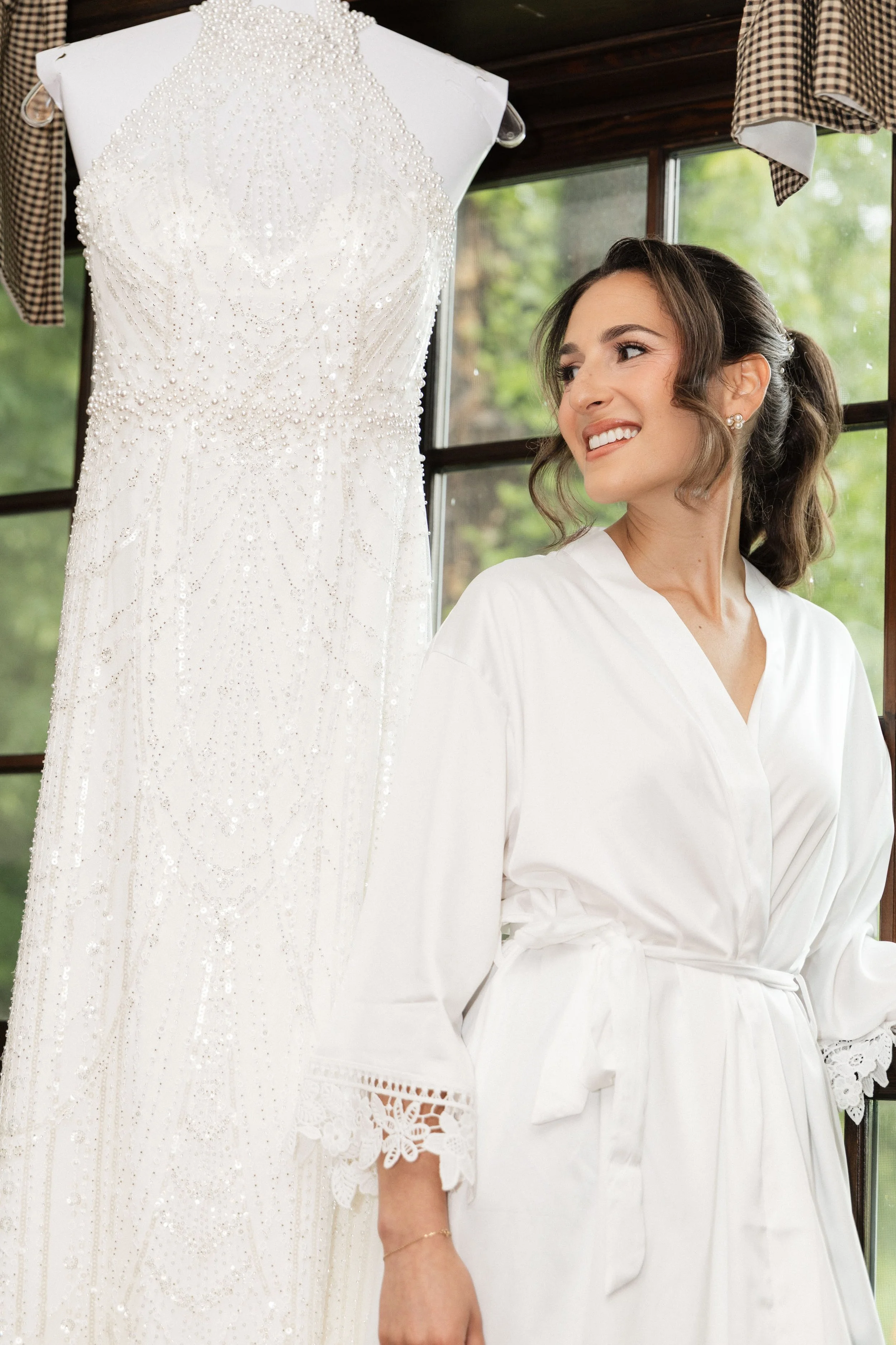 A woman is smiling while looking at a white wedding dress decorated with pearls and beads. The dress is on a mannequin, and she is wearing a white robe with lace cuffs.